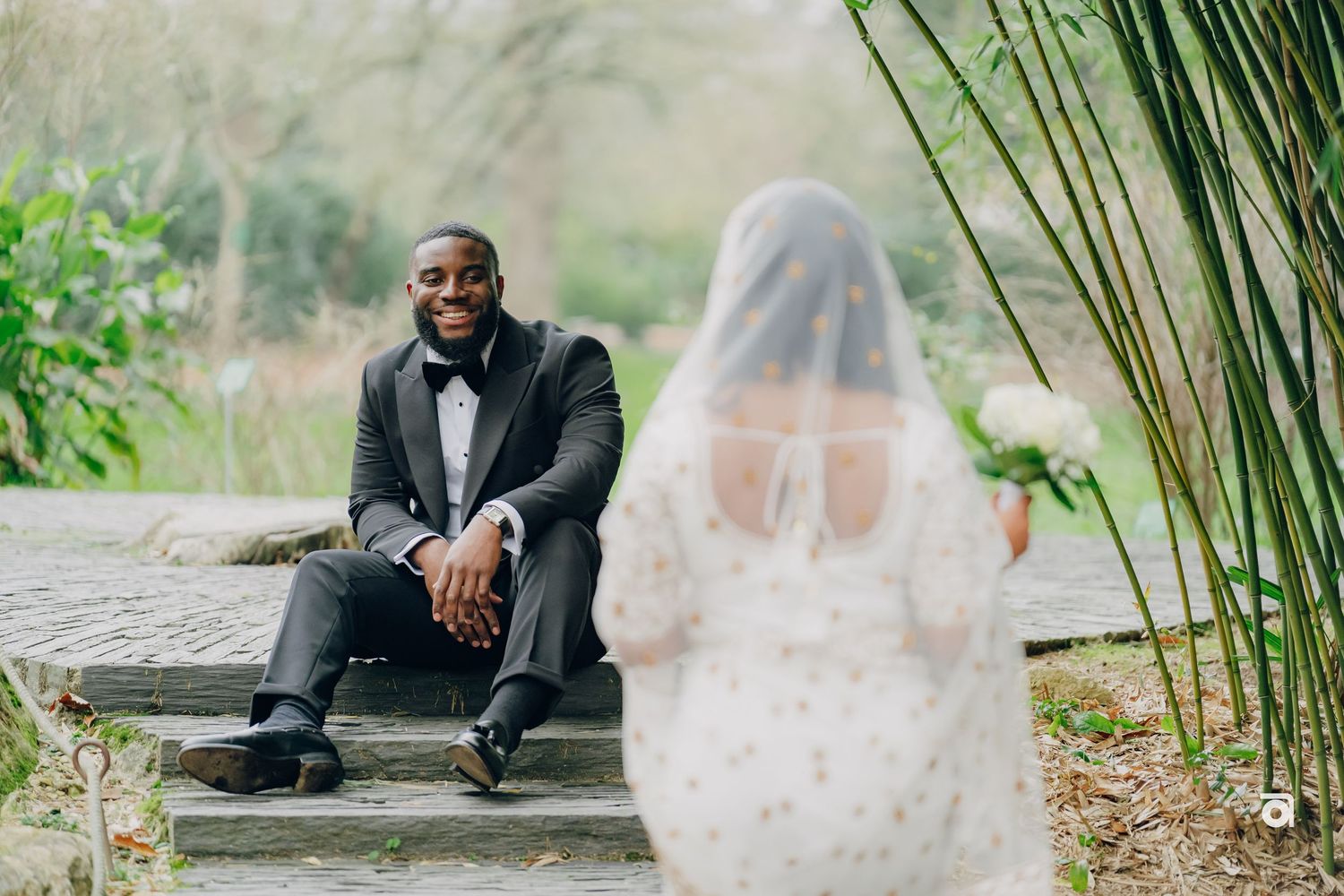 Elegant wedding scene in a garden setting with grey suit and flowing white veil among lush greenery.