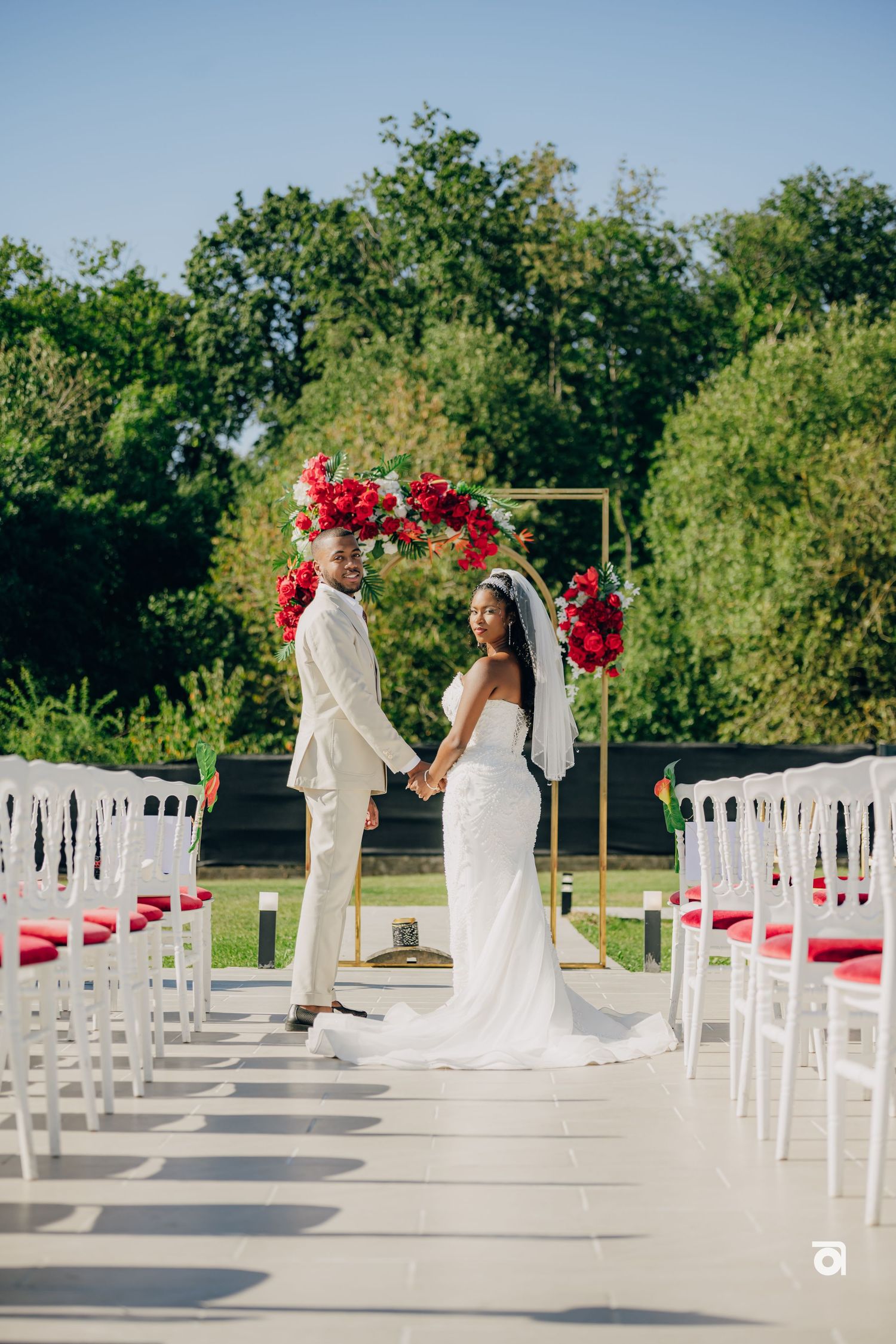 A couple stands under a red floral arch at an outdoor wedding ceremony with white chairs lining the aisle.