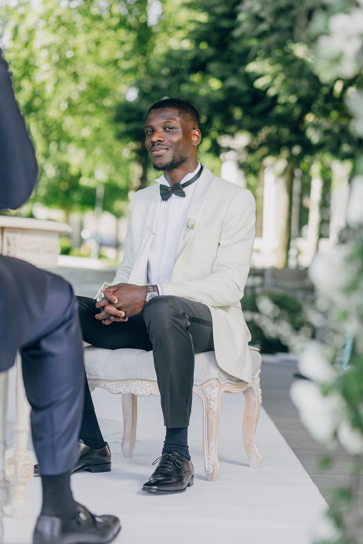 A guest in a white tuxedo jacket and bowtie sits elegantly on a white chair during an outdoor event.