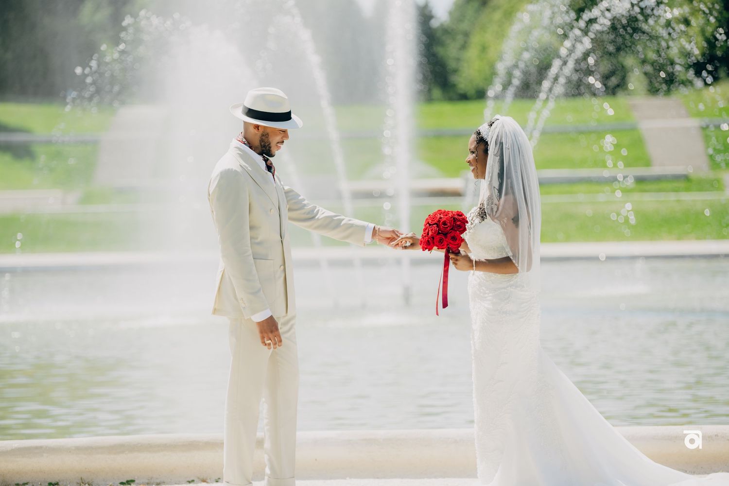 A wedding couple poses near a fountain in a romantic garden setting, with the bride holding a red bouquet.