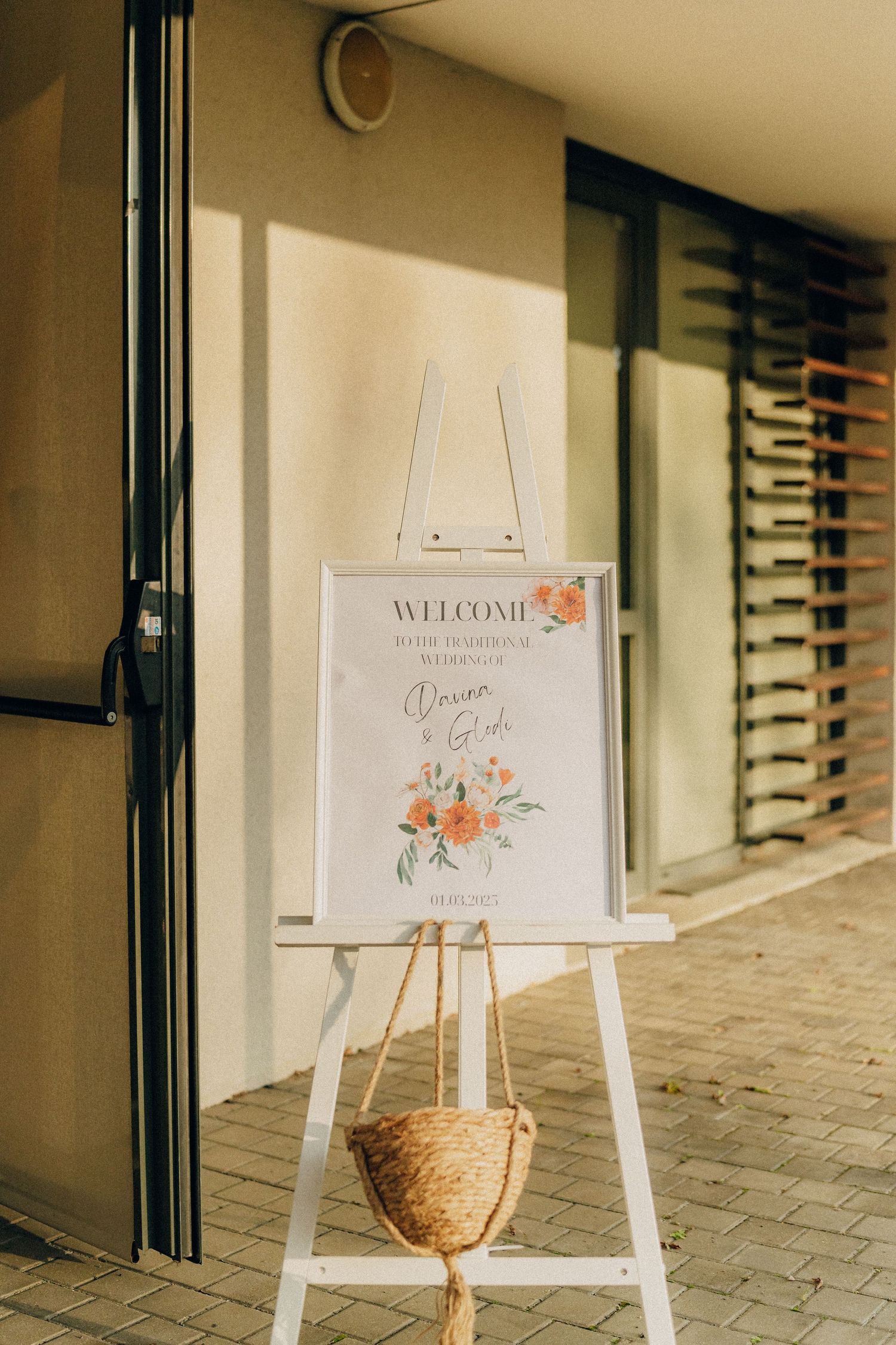 A white welcome sign with floral decorations stands on an easel by a doorway with a woven basket underneath.