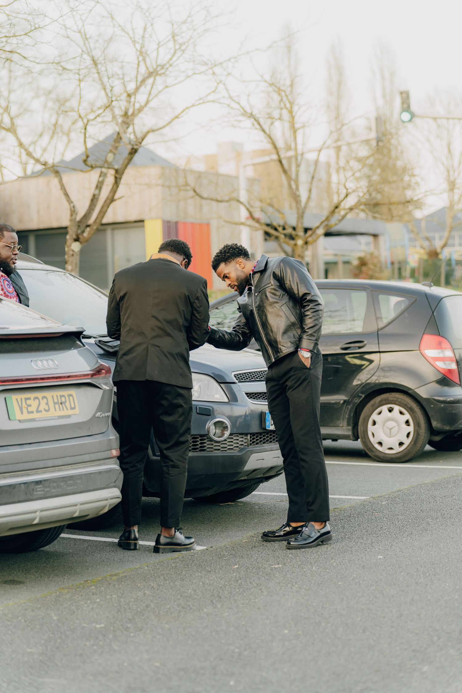 Three people gathered around the back of parked cars in an urban area on an overcast day.