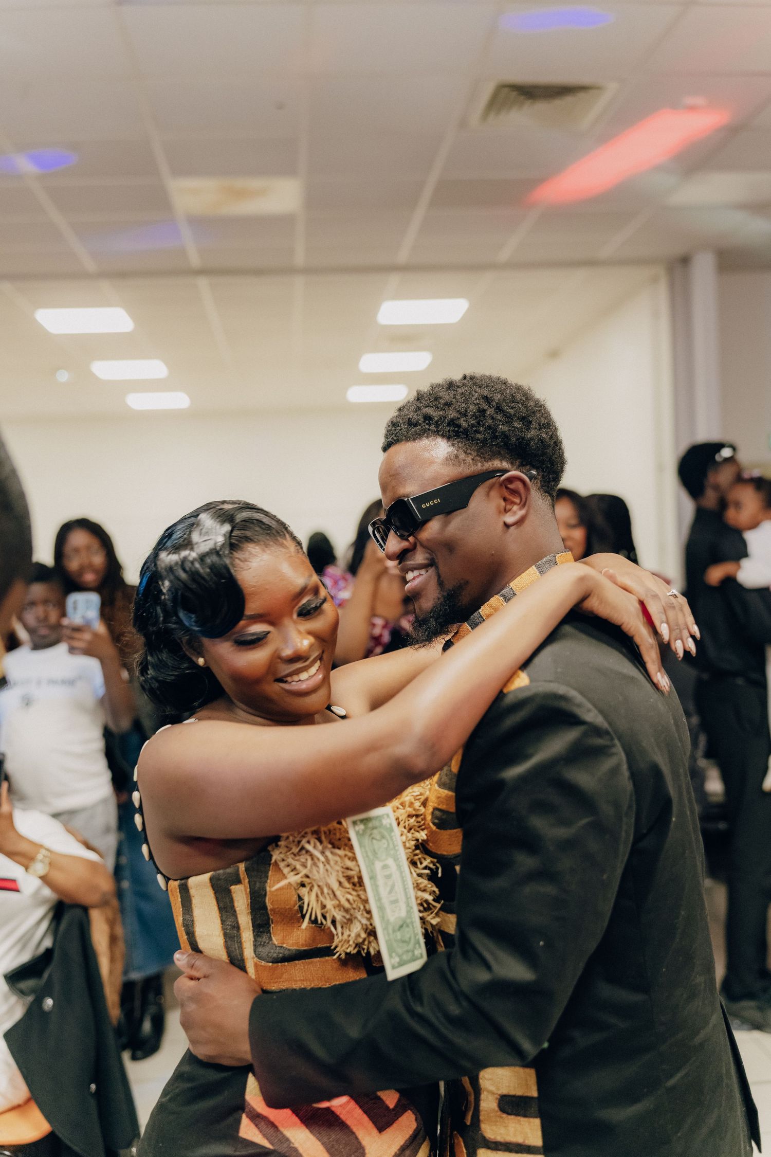 A joyful couple shares an intimate moment at an indoor celebration while guests look on in the background.