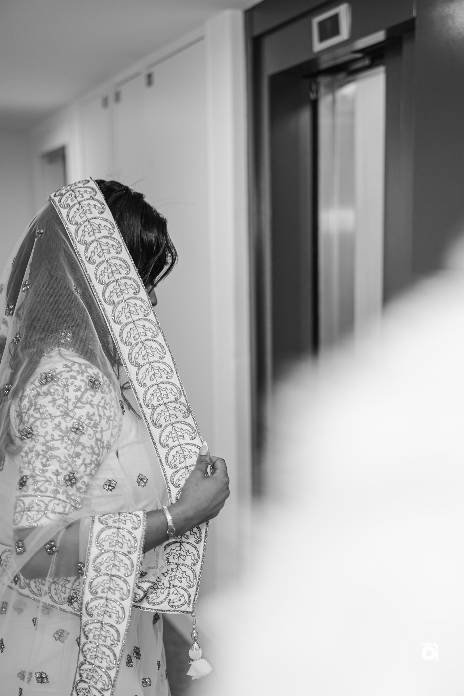 Side view of a bride in an ornate white sari with detailed embroidery standing near an elevator.