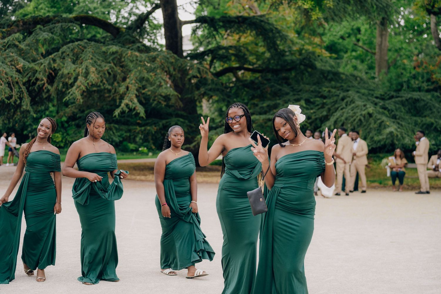 Bridesmaids in elegant emerald green dresses pose together on a path surrounded by lush trees at an outdoor wedding.