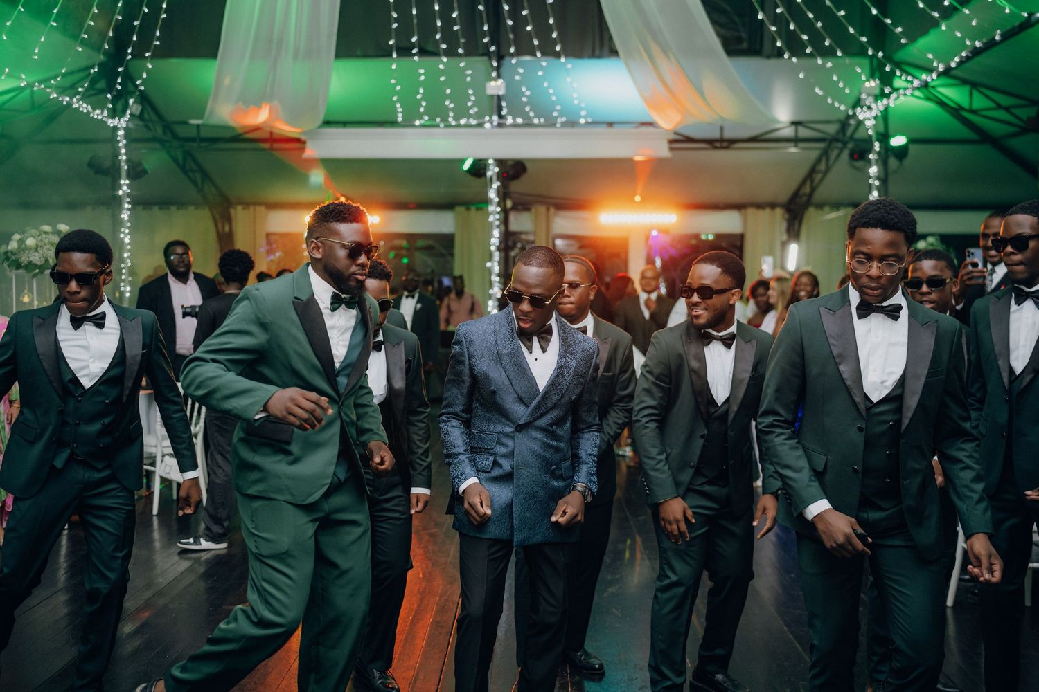 Groomsmen in formal suits dance and celebrate at a wedding reception under green and orange lighting.