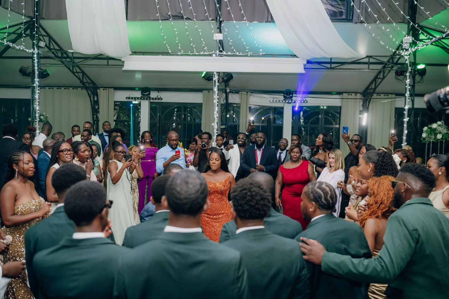 Large group of wedding guests celebrating on a packed dance floor with colorful lighting.