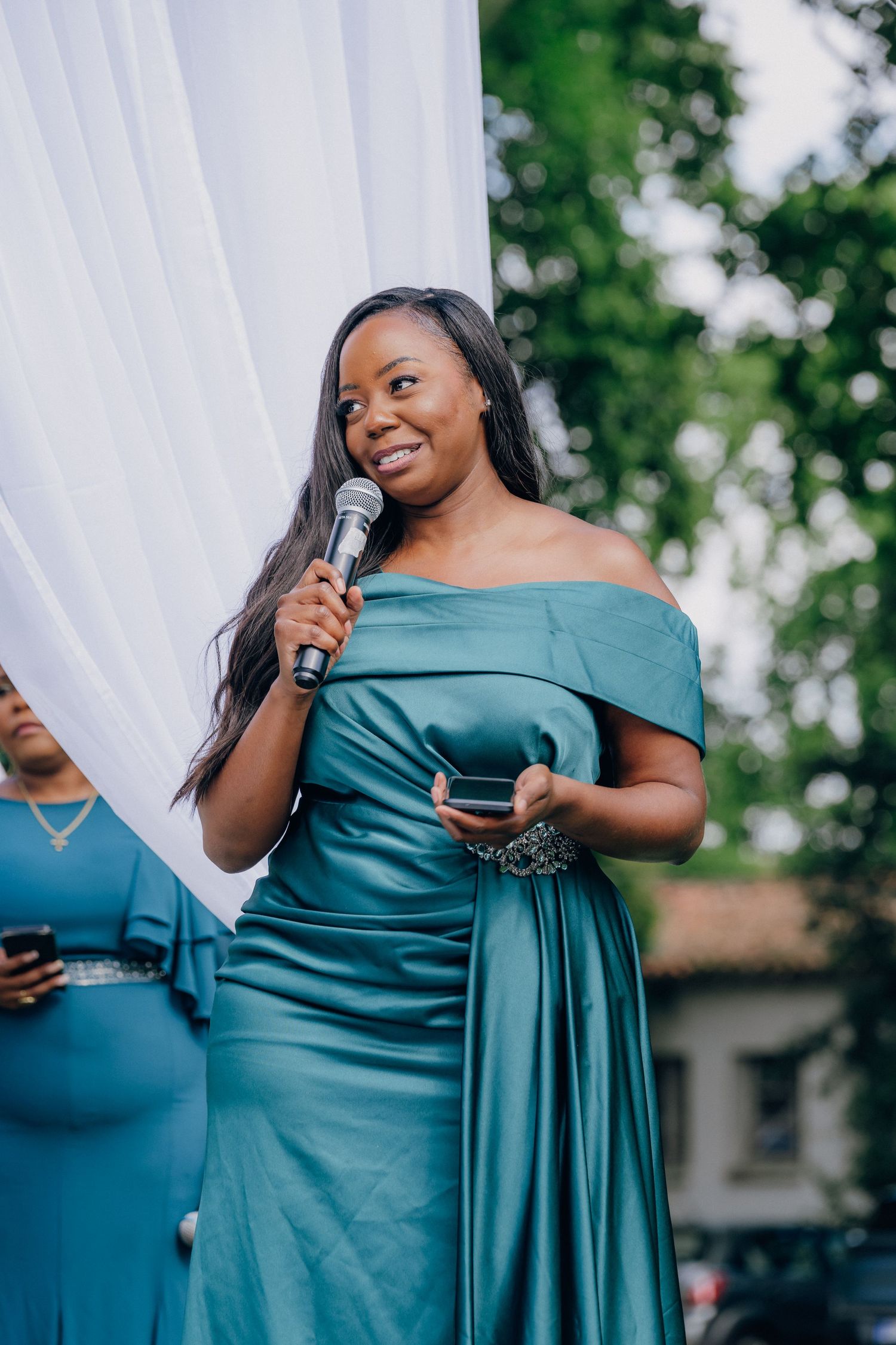 Person in teal off-shoulder dress speaking into microphone at outdoor event with white draping.