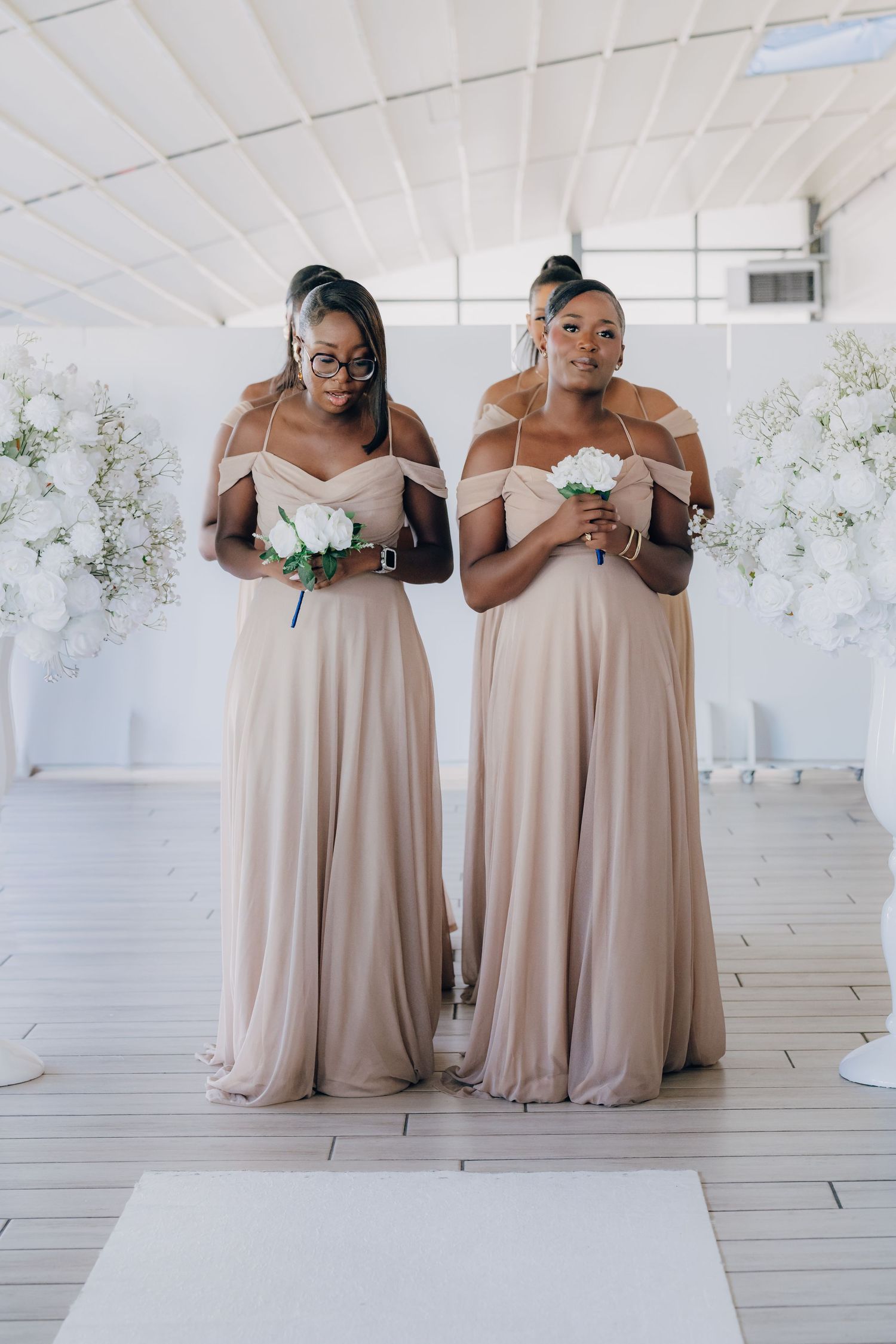 Bridesmaids in champagne-colored floor length dresses pose together in an elegant white room with floral decor.
