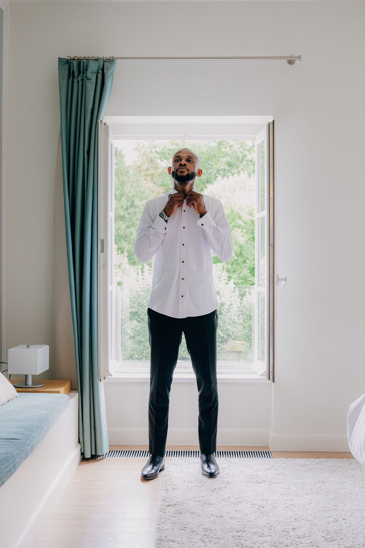 A person in a white shirt and dark pants stands by a window in an empty room with teal curtains.