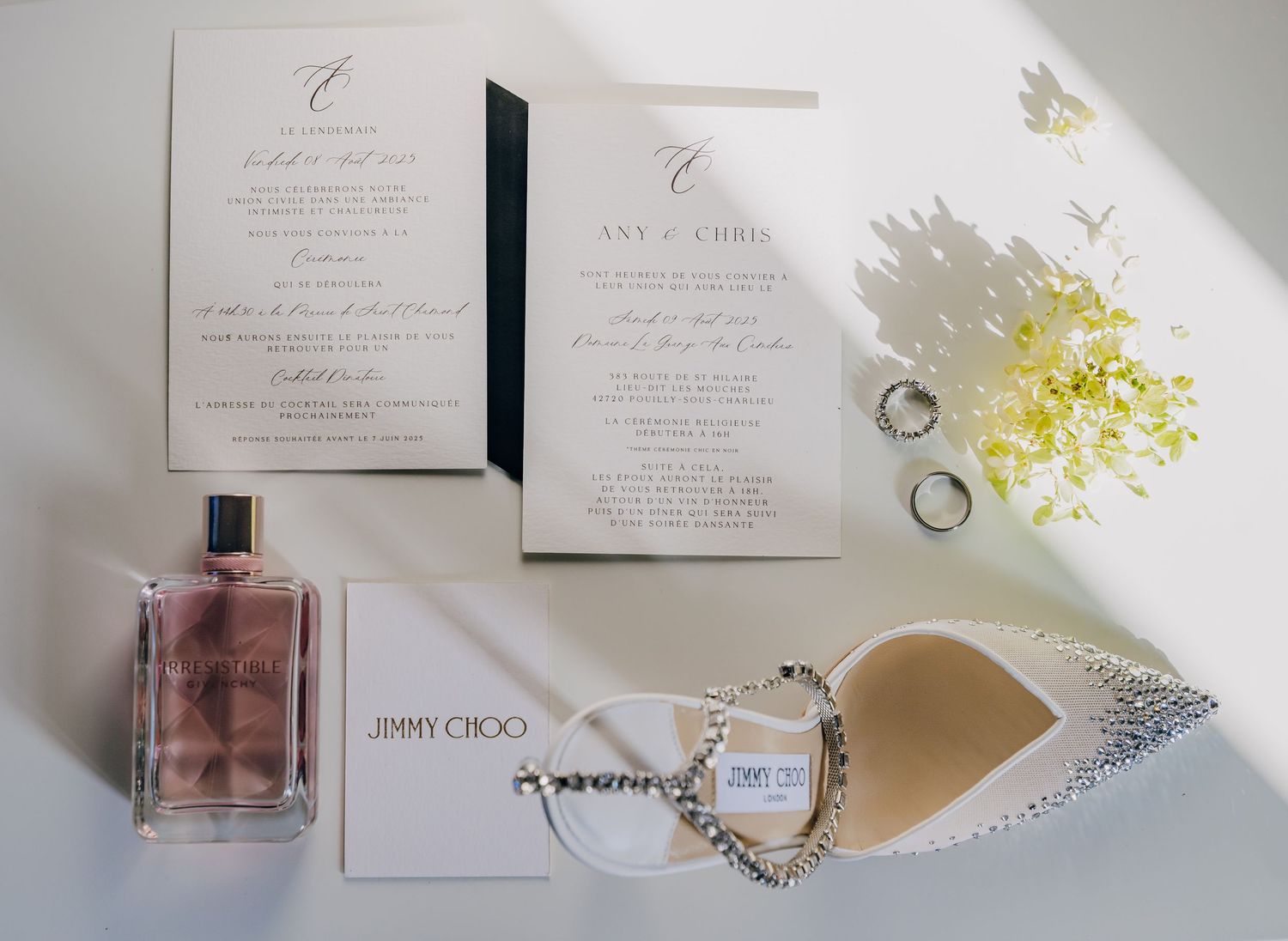 Wedding invitations, perfume bottle, and rings arranged on white surface with natural lighting.