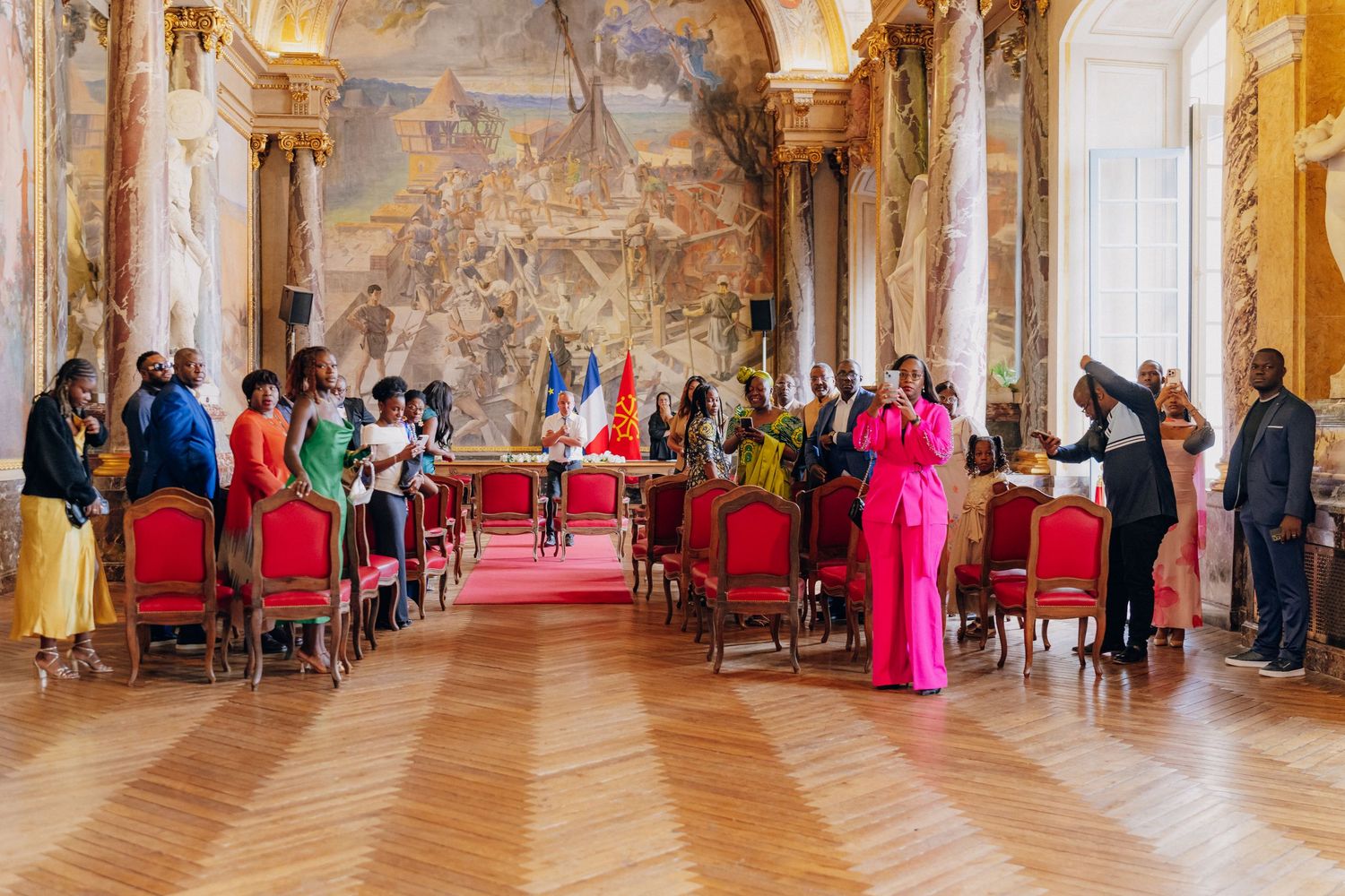 A civil ceremony takes place in an ornate palace room with frescoed walls, red chairs, and guests in colorful attire.