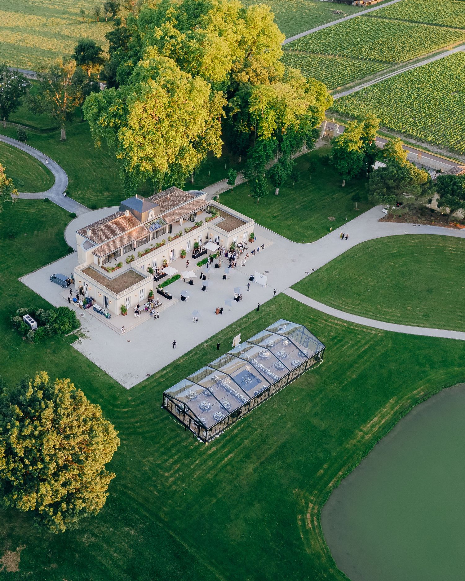 Aerial view of a elegant winery estate with manicured lawns, vineyard rows, greenhouse, and pond at golden hour.