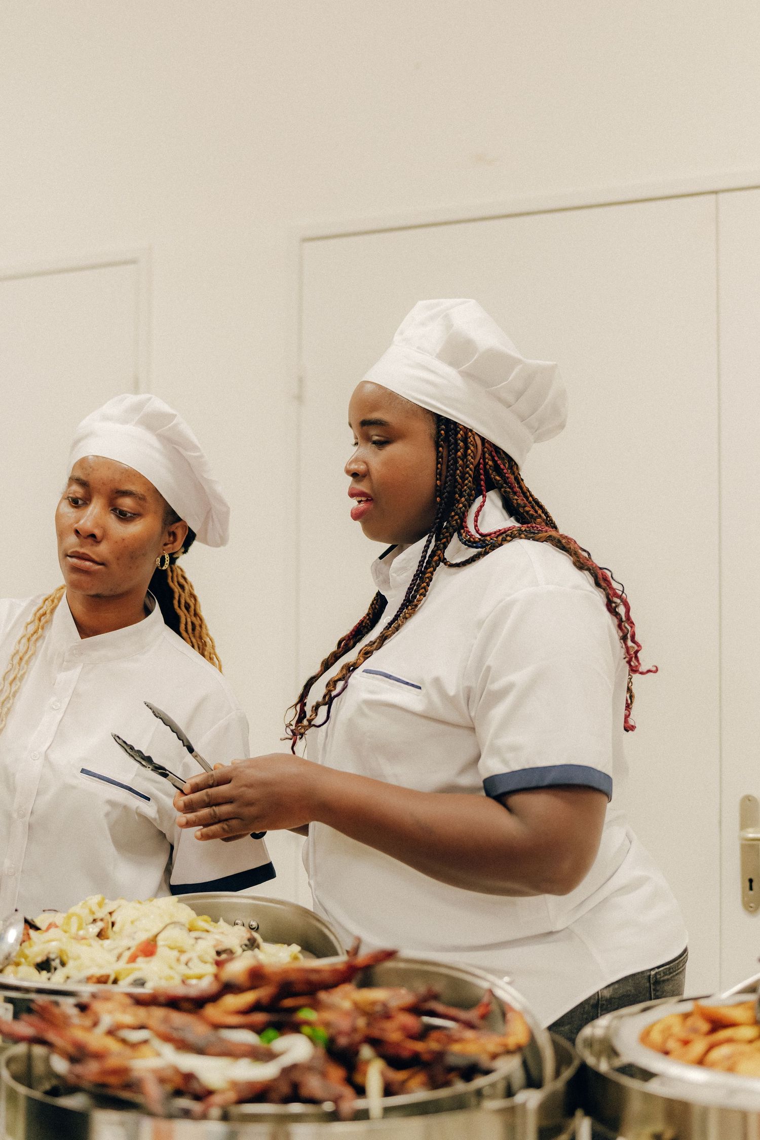 Two chefs in white uniforms and chef hats work together serving food at a catered buffet event.