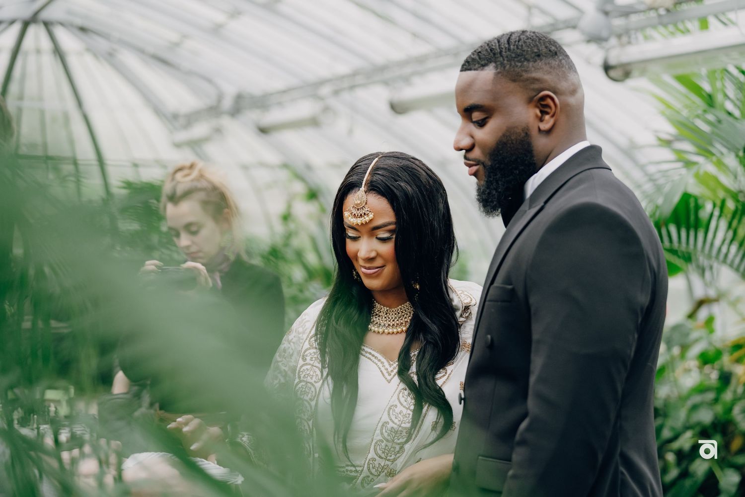An intimate wedding ceremony moment inside a greenhouse with a couple in elegant attire surrounded by tropical plants.