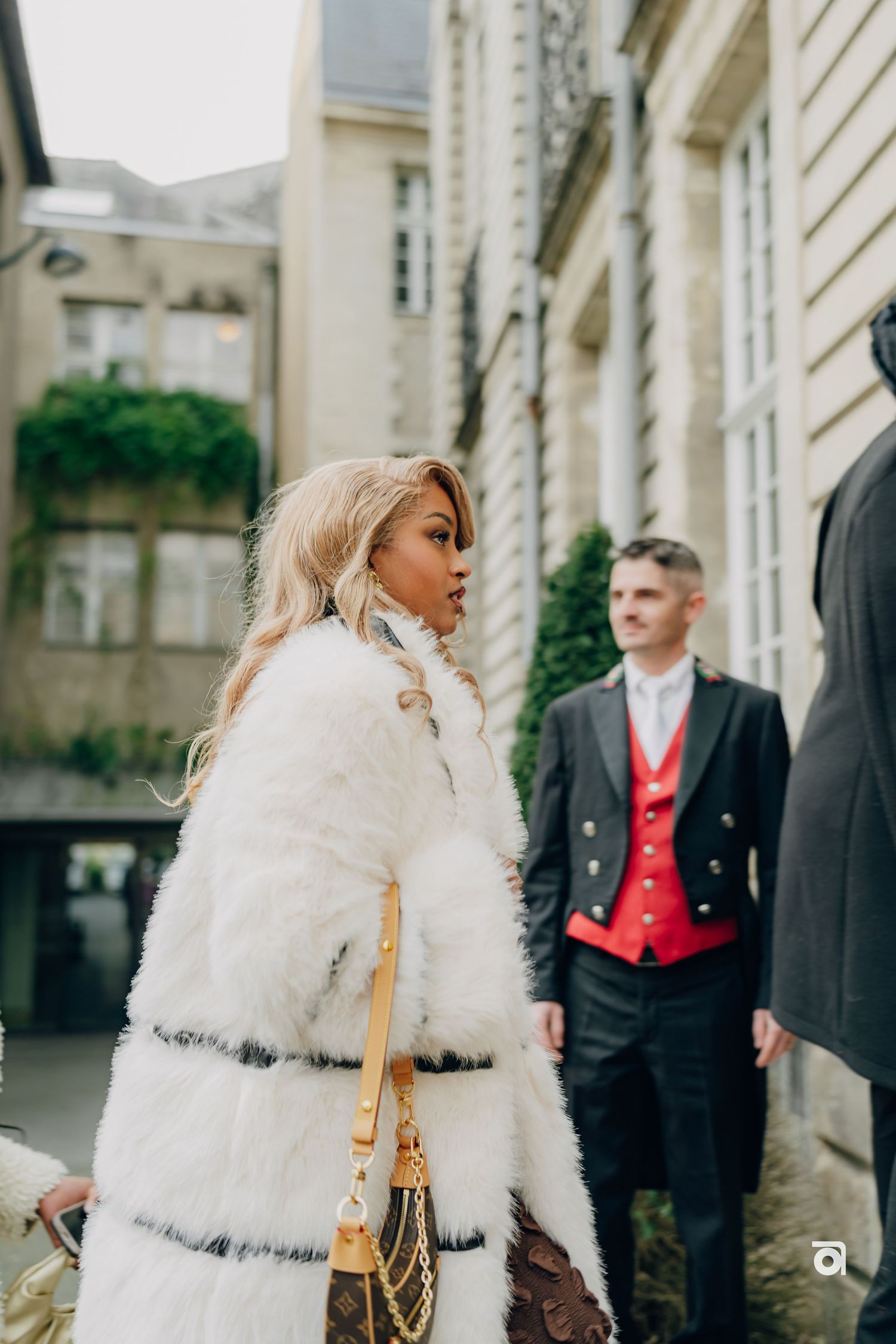 A couple in formal attire stands outside a building, with someone wearing a luxurious white fur coat and holding a gold purse.