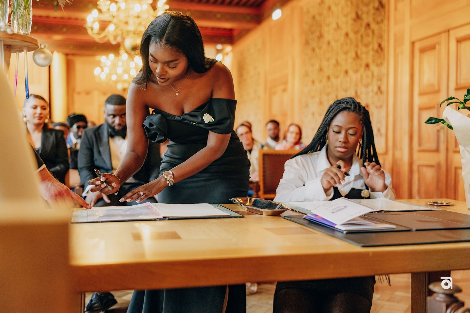 Wedding ceremony attendees signing official documents at a wooden table in an elegant venue.