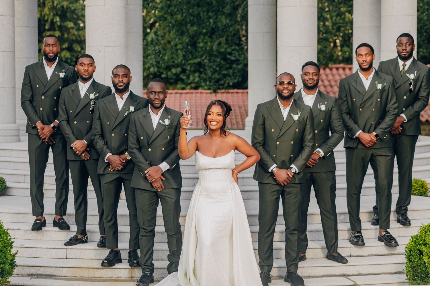 Wedding party in gray suits poses with member in white dress against columned backdrop.