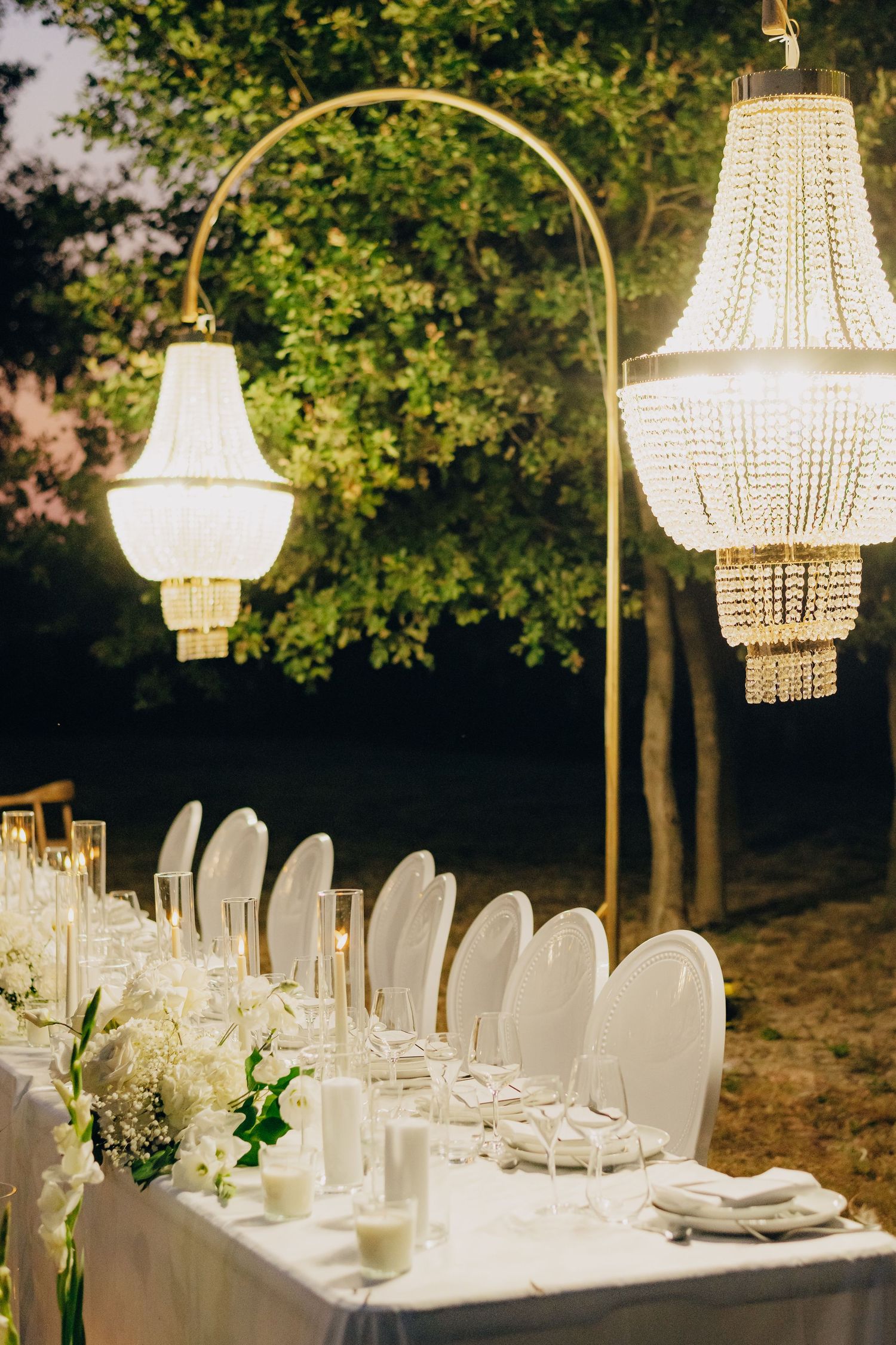 Elegant wedding table setting with crystal chandeliers and white chairs under evening sky.