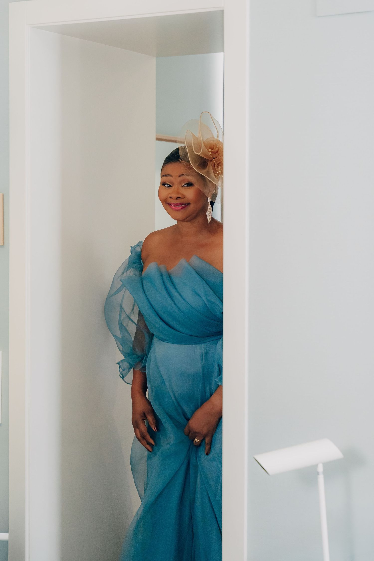 Someone wearing an elegant blue off-shoulder dress poses in a white hallway.