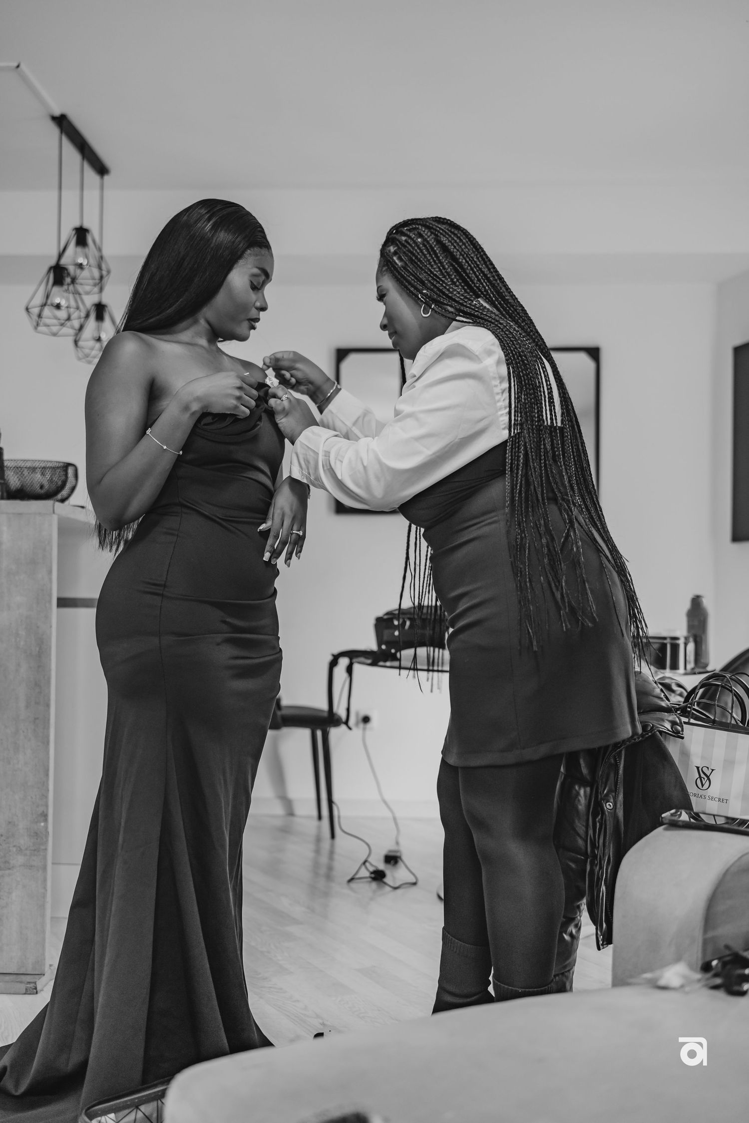 A person in a fitted black gown gets assistance with final dress preparations in an intimate black and white moment.