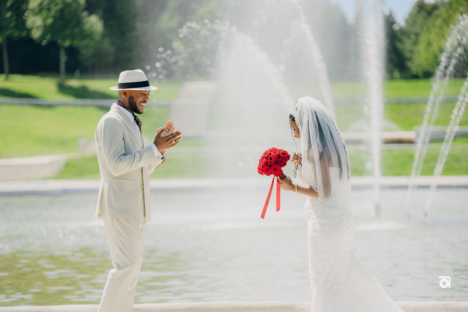 A wedding ceremony taking place in front of a fountain with white dress and suit against a sunny garden backdrop.
