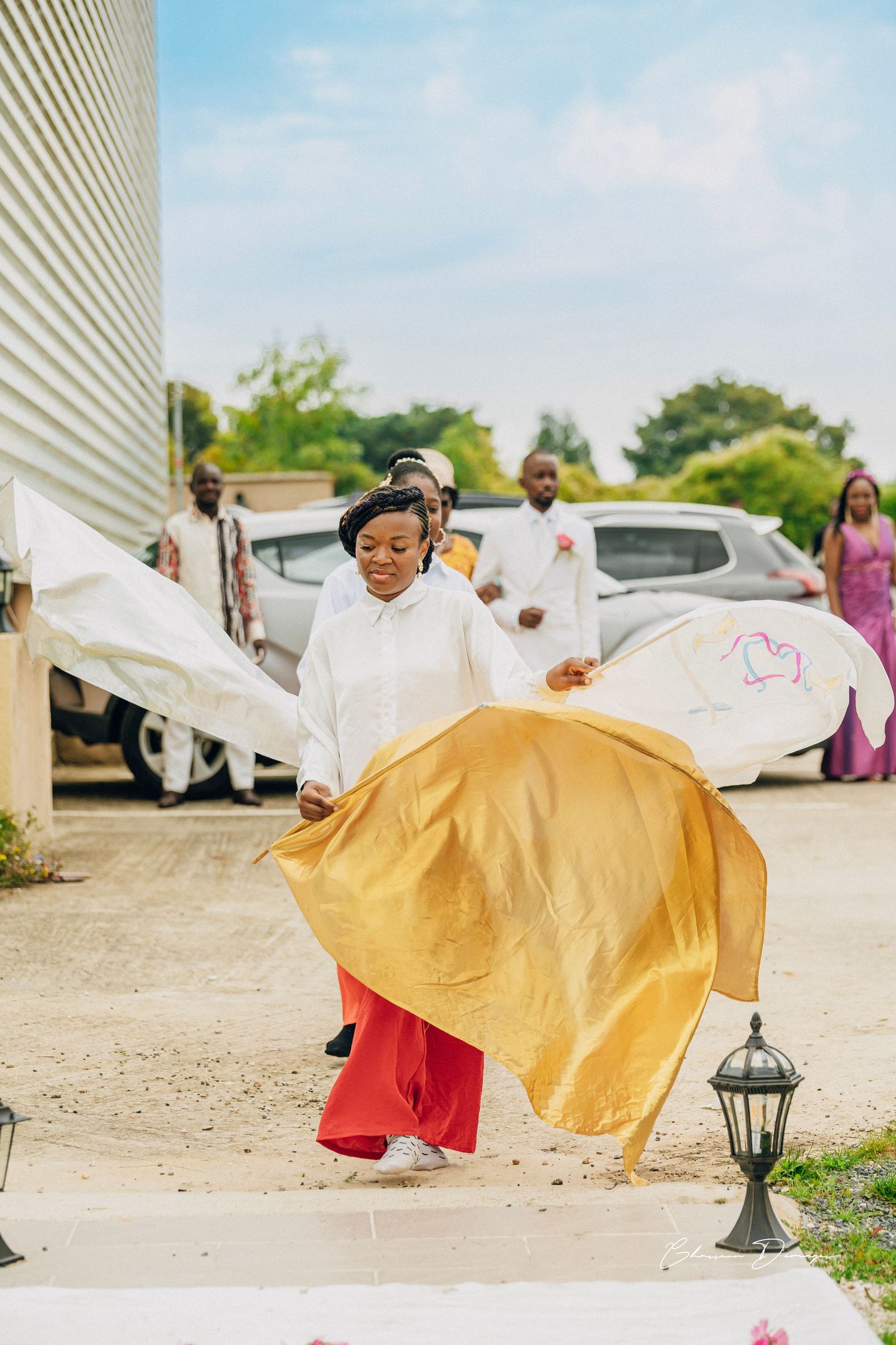 A young attendant in white twirls a golden fabric outside during wedding festivities.