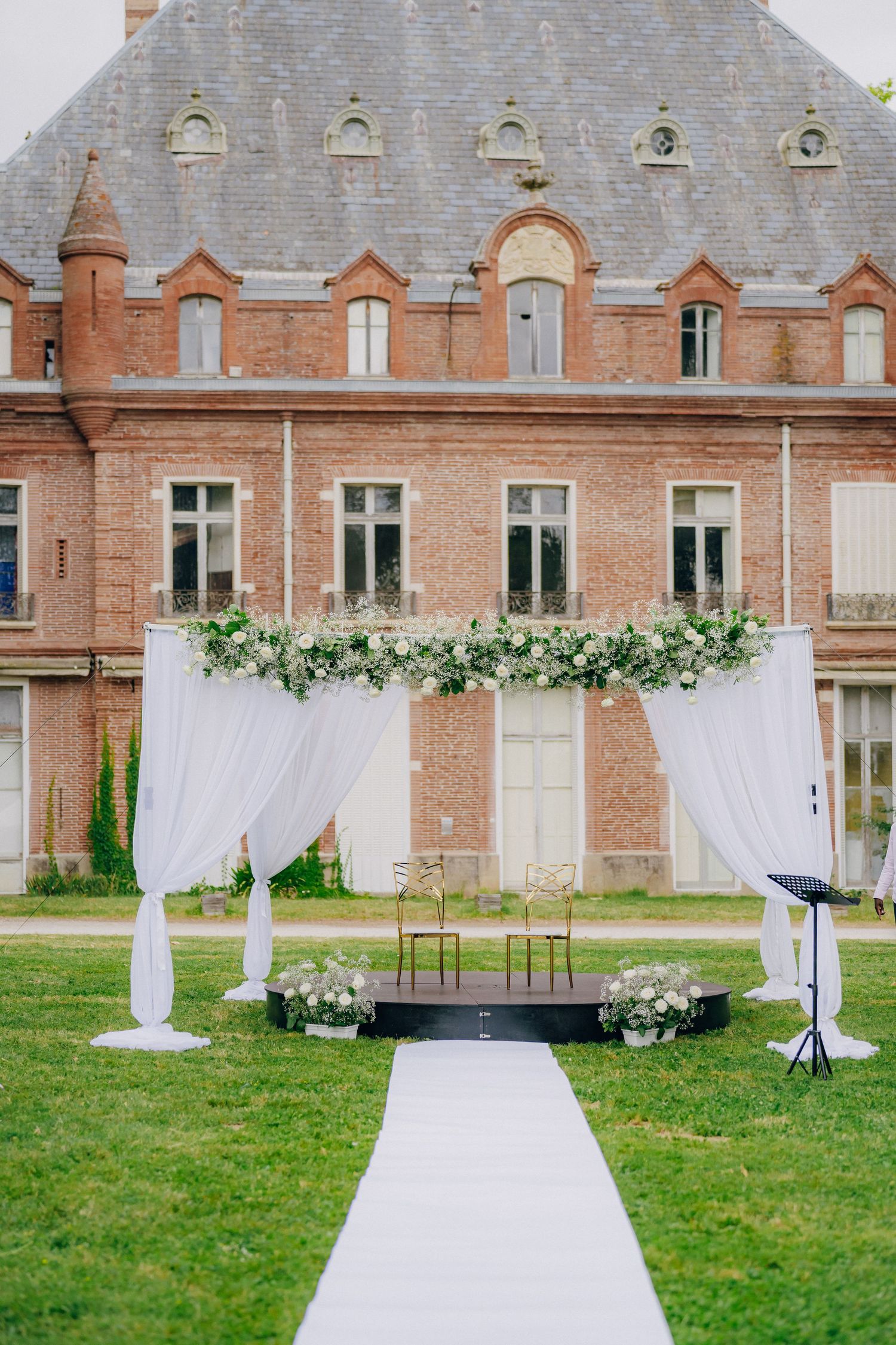 Elegant outdoor wedding ceremony setup with white draped fabric and floral arrangements in front of a brick mansion.