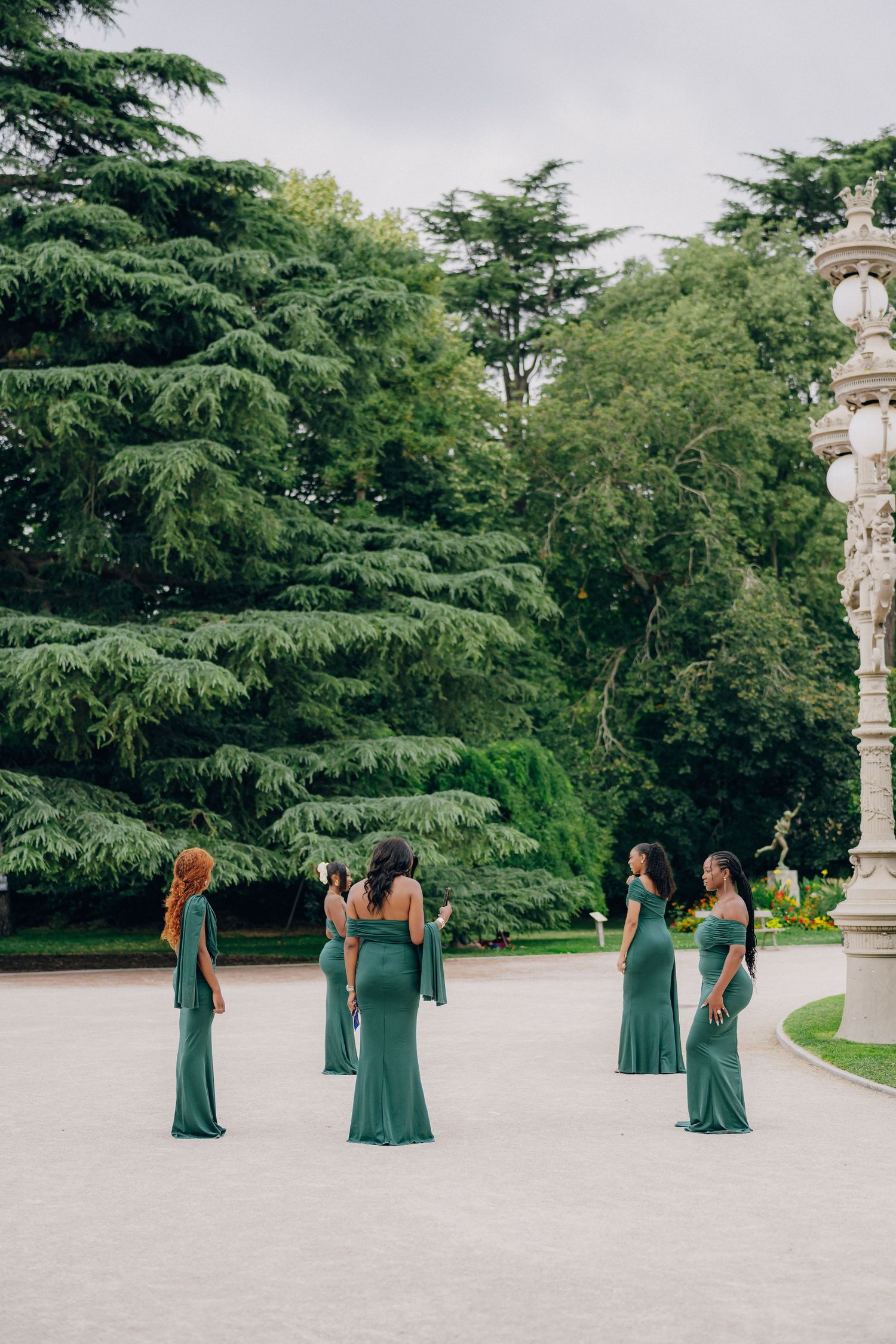 Bridesmaids in matching teal floor-length dresses gather on a garden path near evergreen trees and ornate architecture.