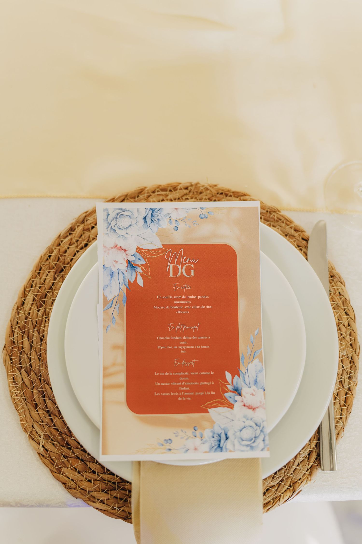 Wedding menu card with orange and blue floral design placed on white plate with woven placemat.