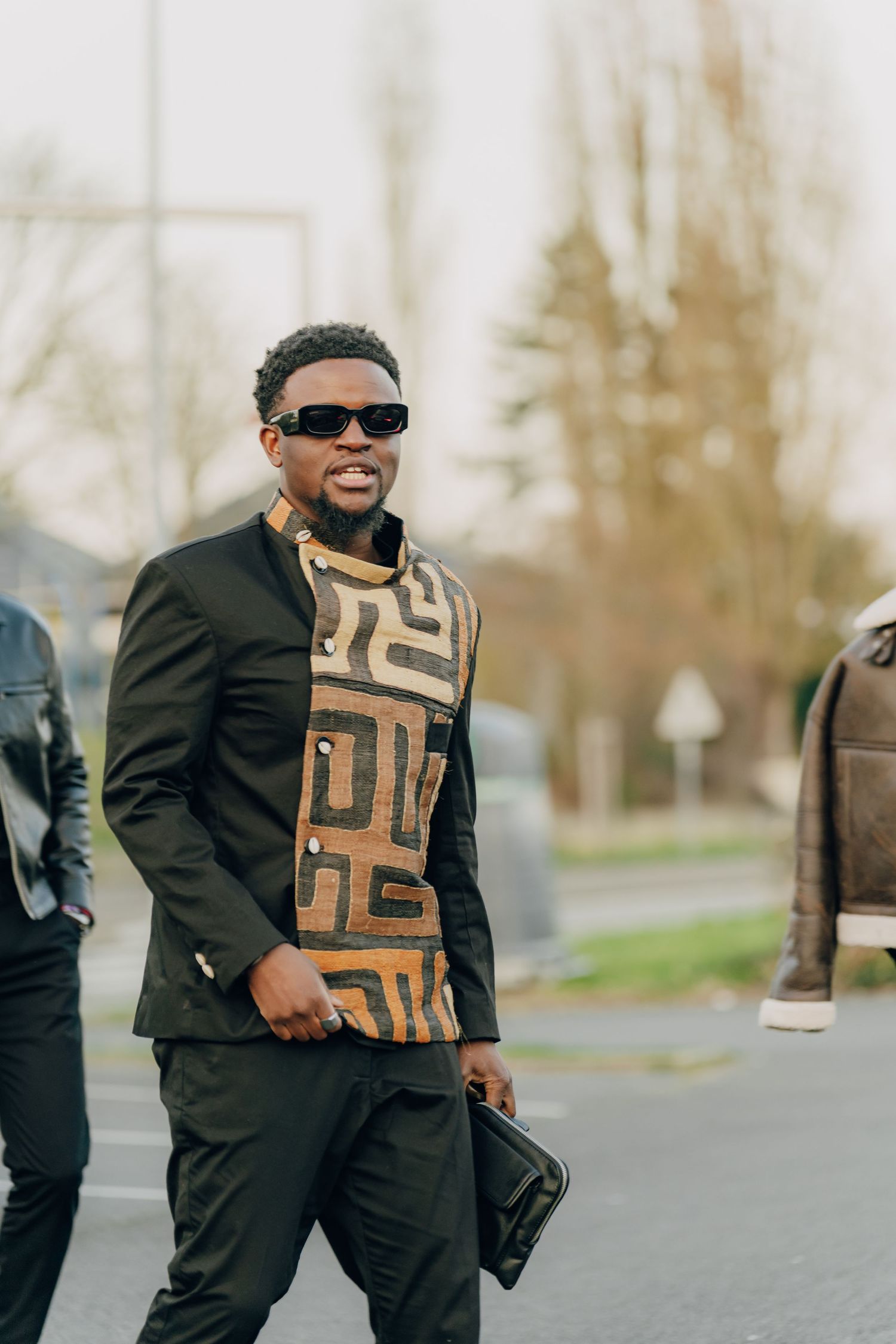 Stylish figure in black suit and geometric patterned scarf walks confidently while wearing sunglasses and carrying a bag.