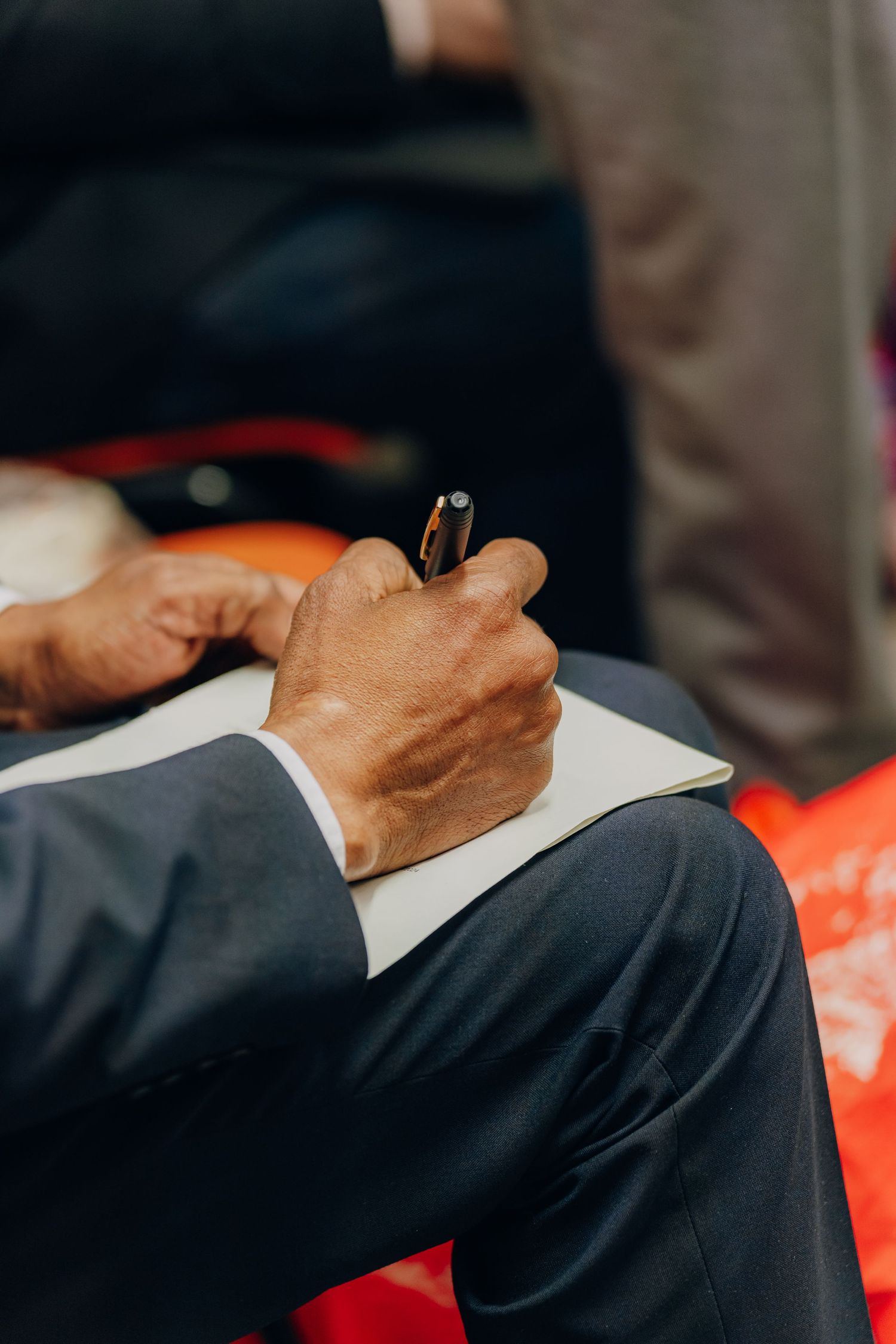 Close-up of hands holding a pen while writing in a notebook on lap wearing formal business attire.