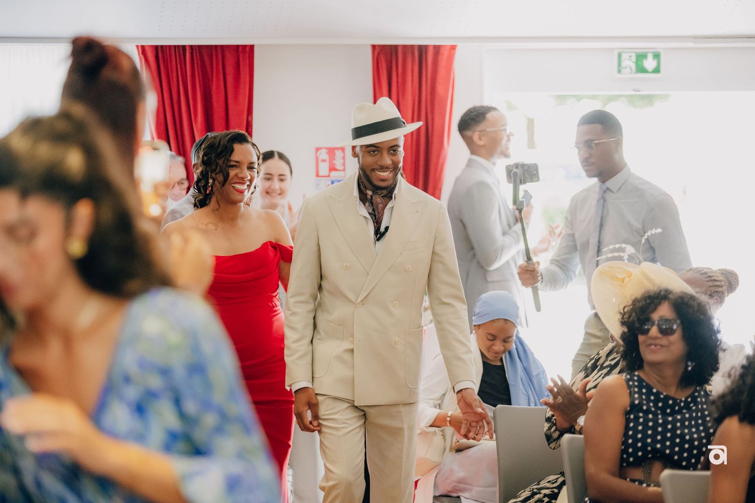 Wedding guests gather in an indoor venue decorated with red curtains as an attendee in a beige suit walks through.