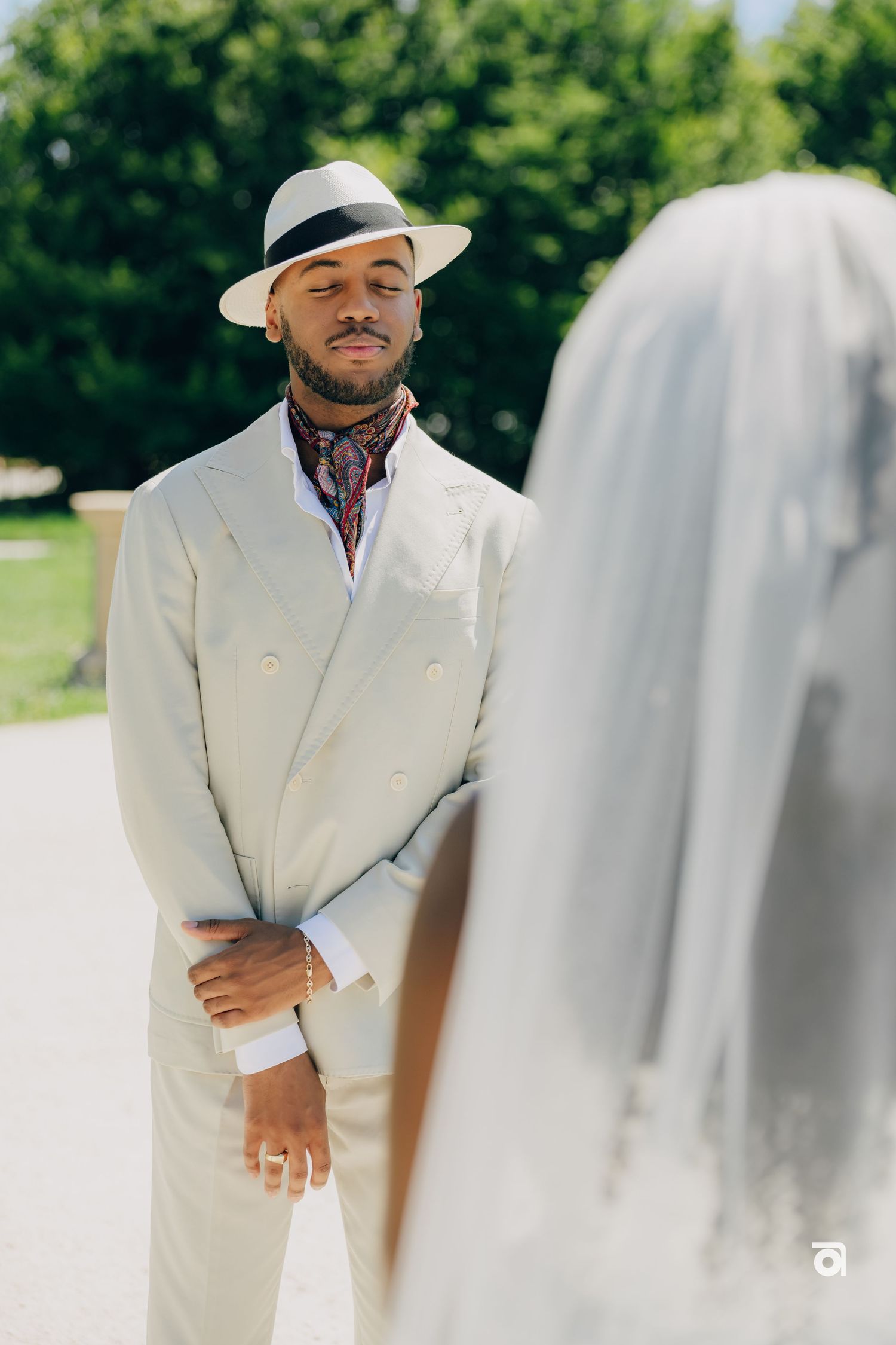 Person in elegant white suit and fedora smiling during an outdoor wedding ceremony.
