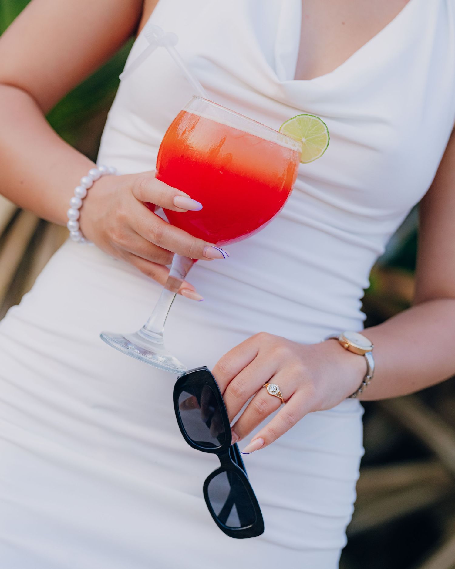 Woman in white dress holding a red cocktail with lime garnish and black sunglasses, wearing pearl bracelet and gold ring.