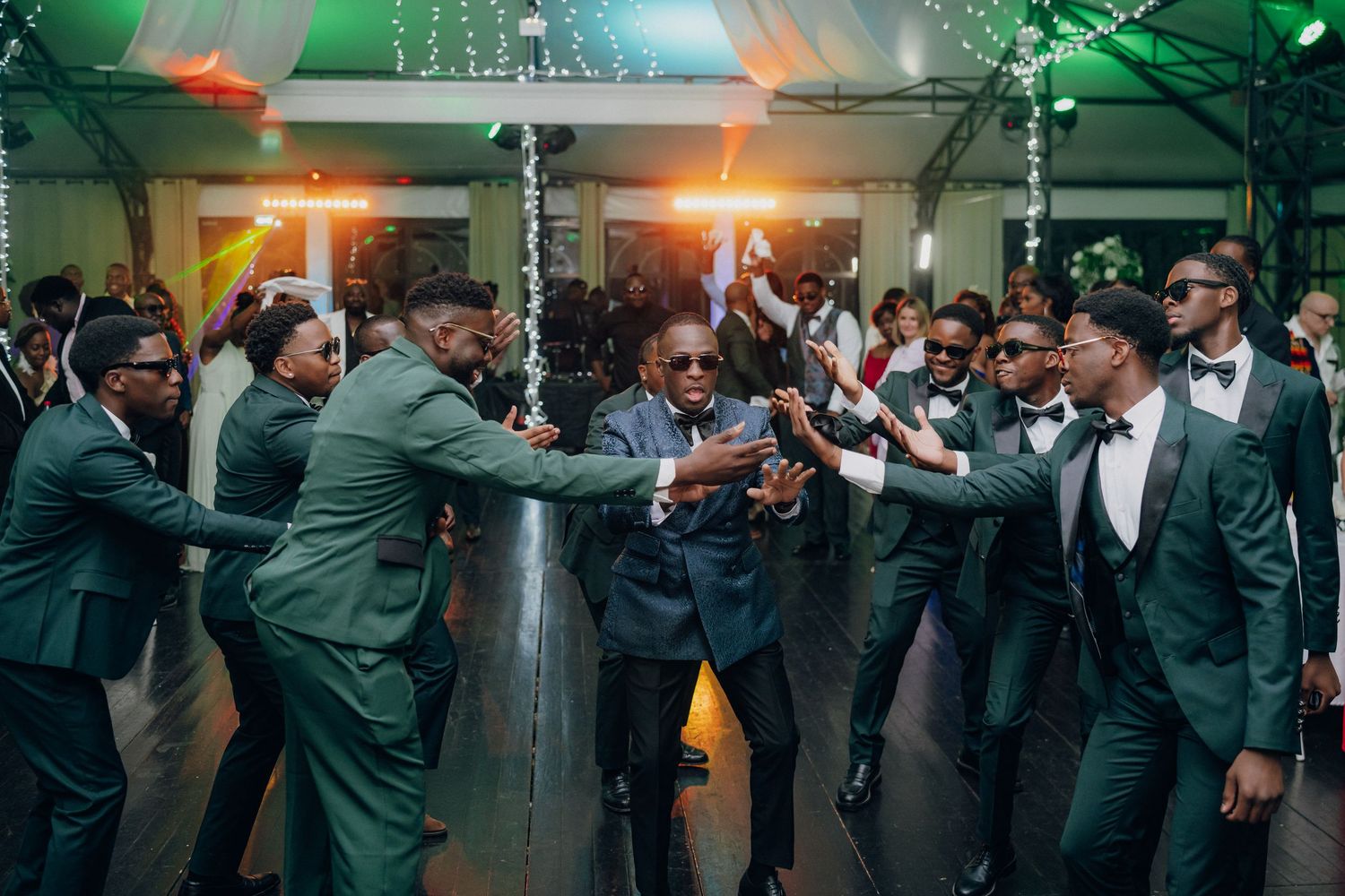 Wedding guests dancing and celebrating on a dance floor illuminated by green and orange lighting effects.