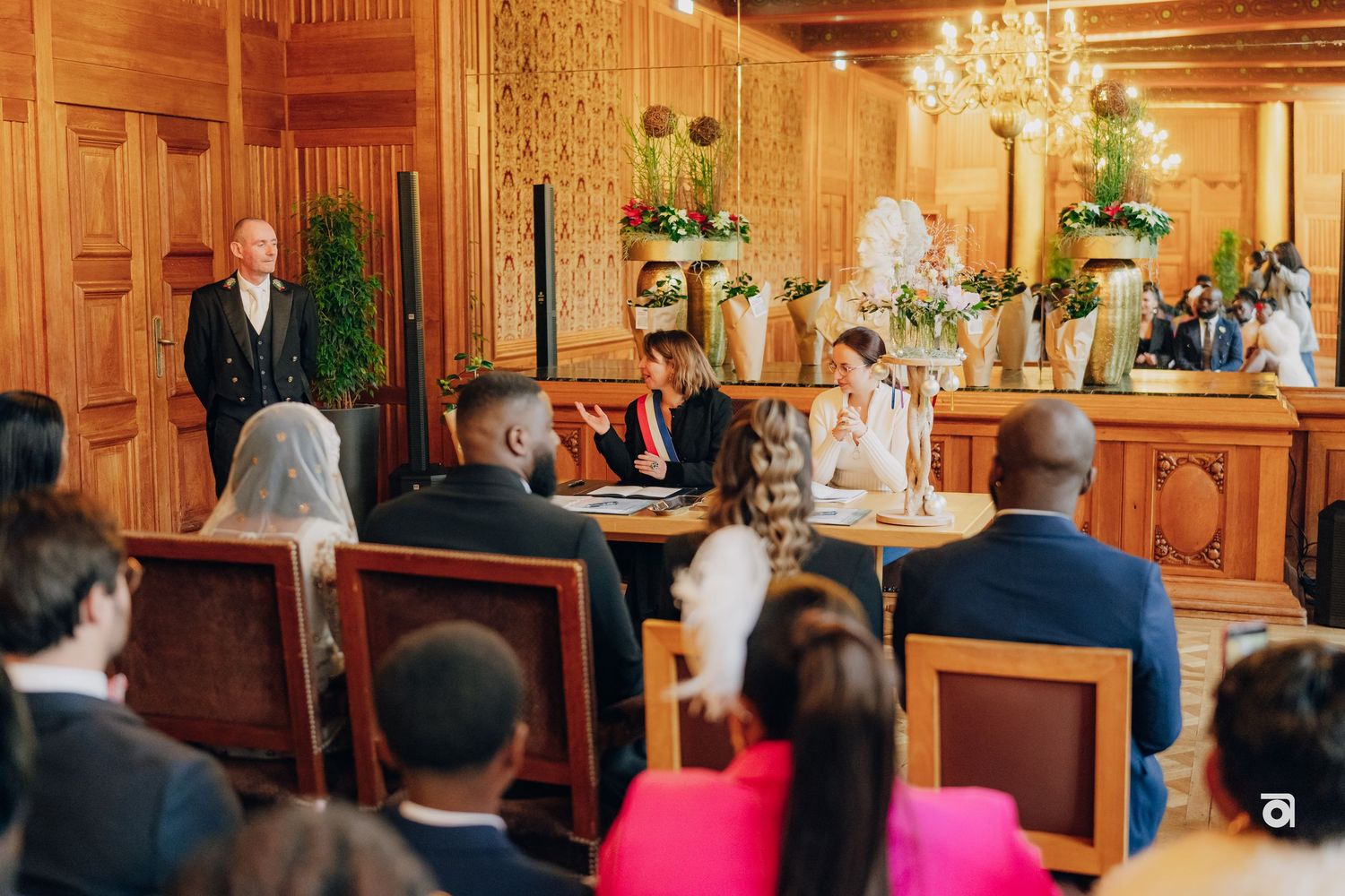 An elegant indoor wedding ceremony takes place in a wood-paneled room with a chandelier and floral arrangements at the altar.
