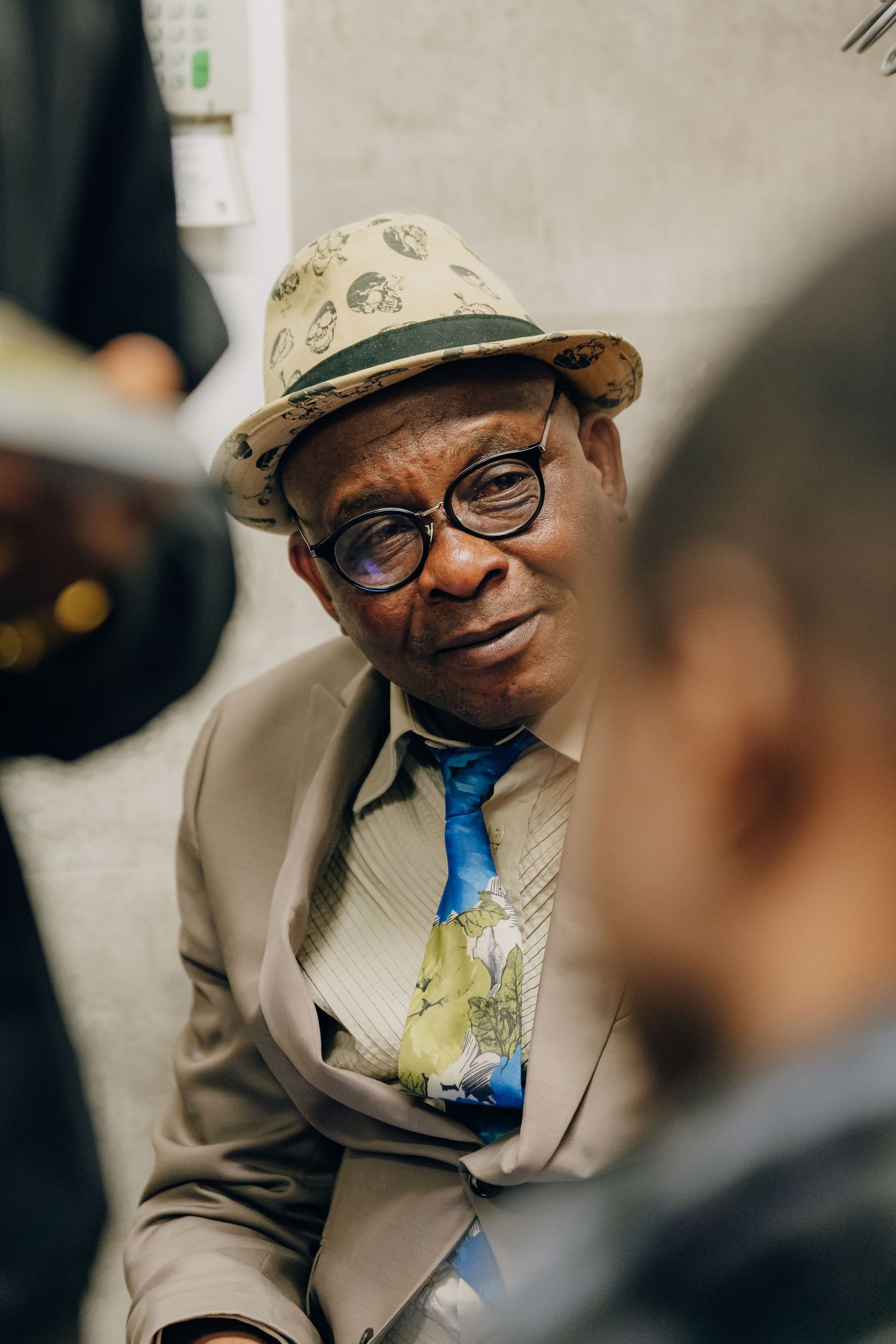 A person wearing a patterned hat, glasses, and blue tie smiles warmly while wearing a light-colored suit jacket.