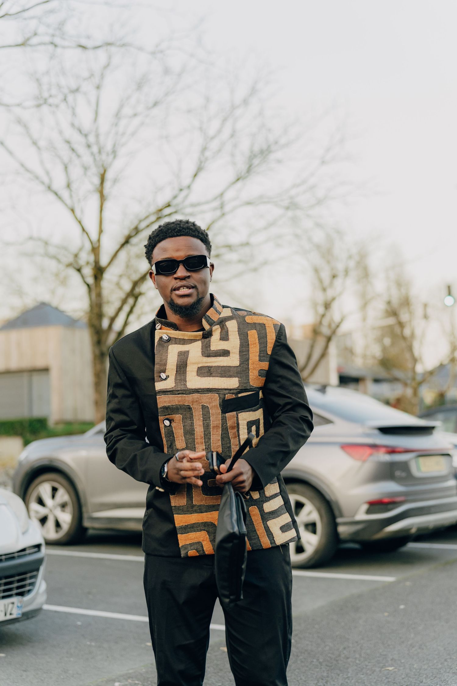 Stylish person wearing sunglasses and black outfit with geometric patterned vest standing in parking lot near cars.