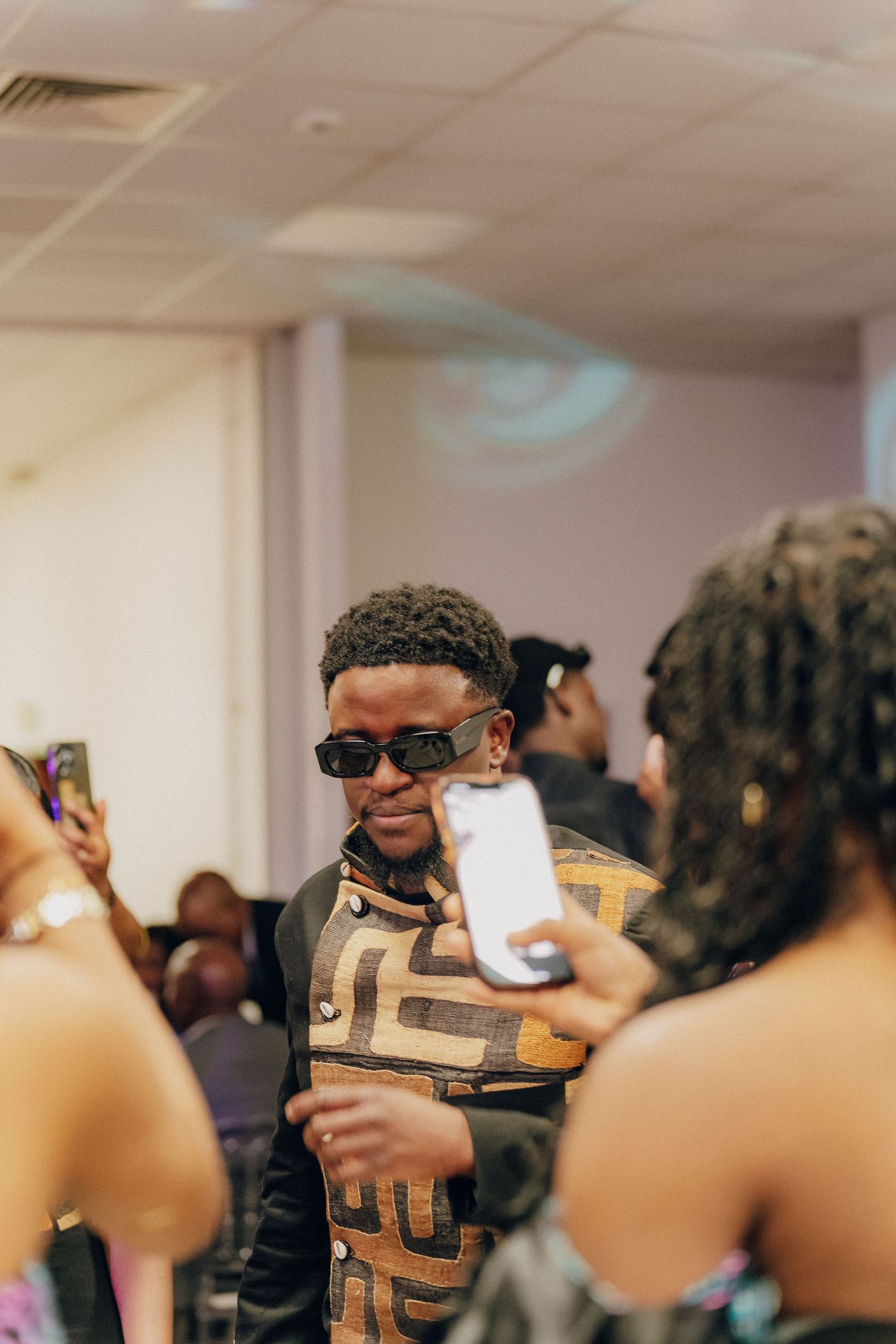 Guest in sunglasses and geometric print shirt poses for photos at indoor event.
