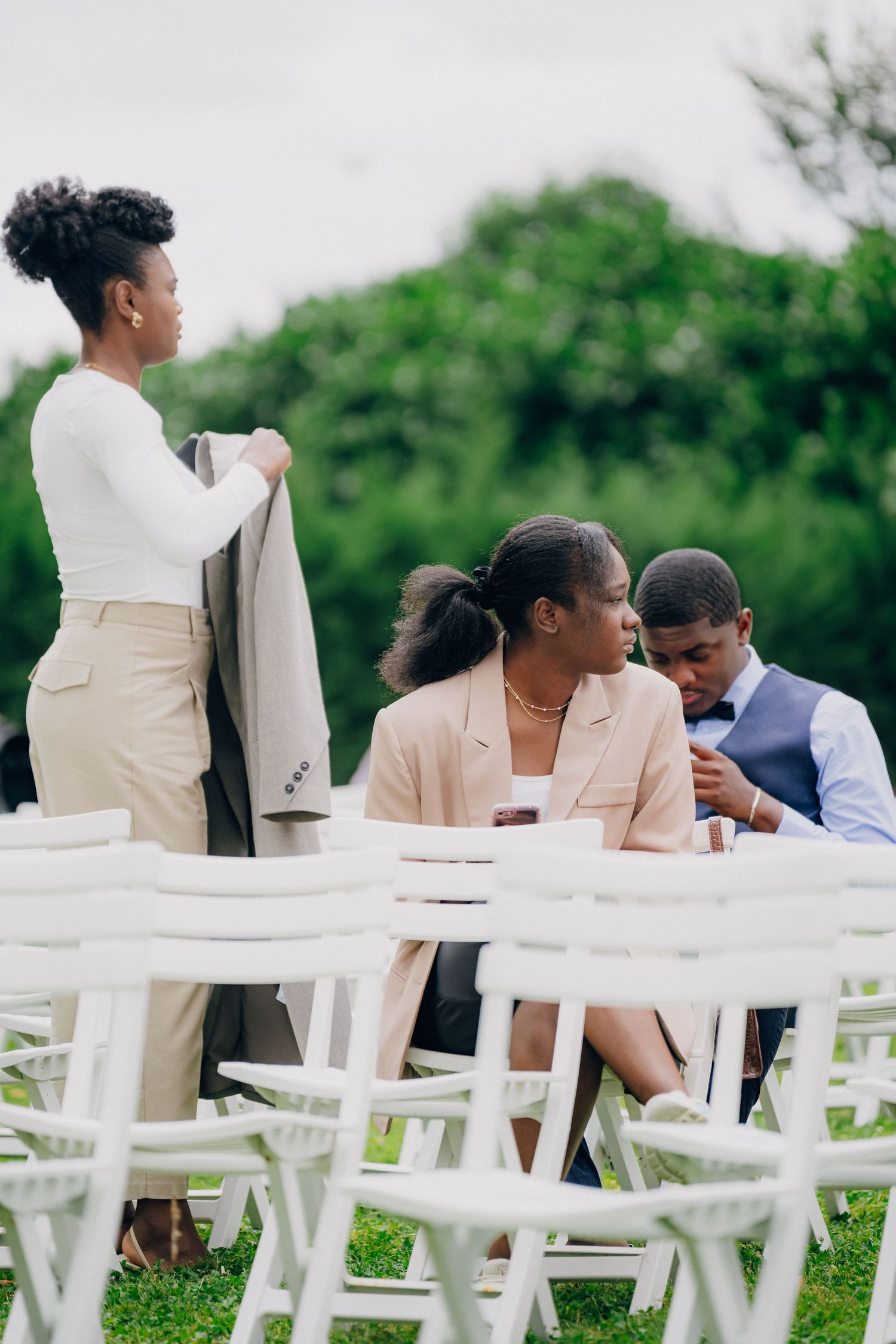 Wedding guests seated in white chairs at an outdoor ceremony with lush greenery in the background.