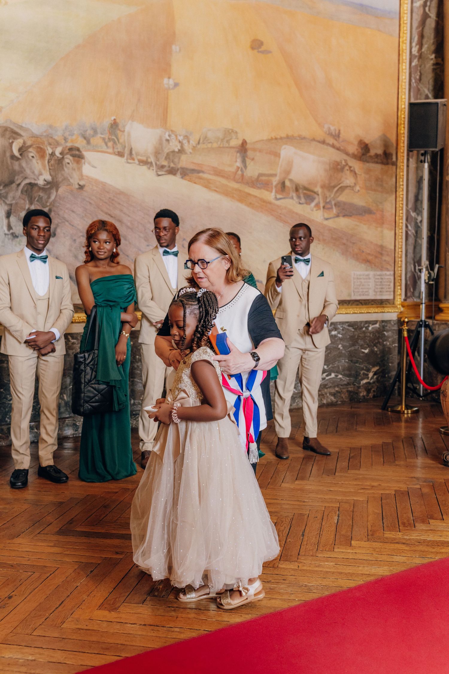 Wedding party gathered in an ornate historic palace room with parquet flooring and red carpet.