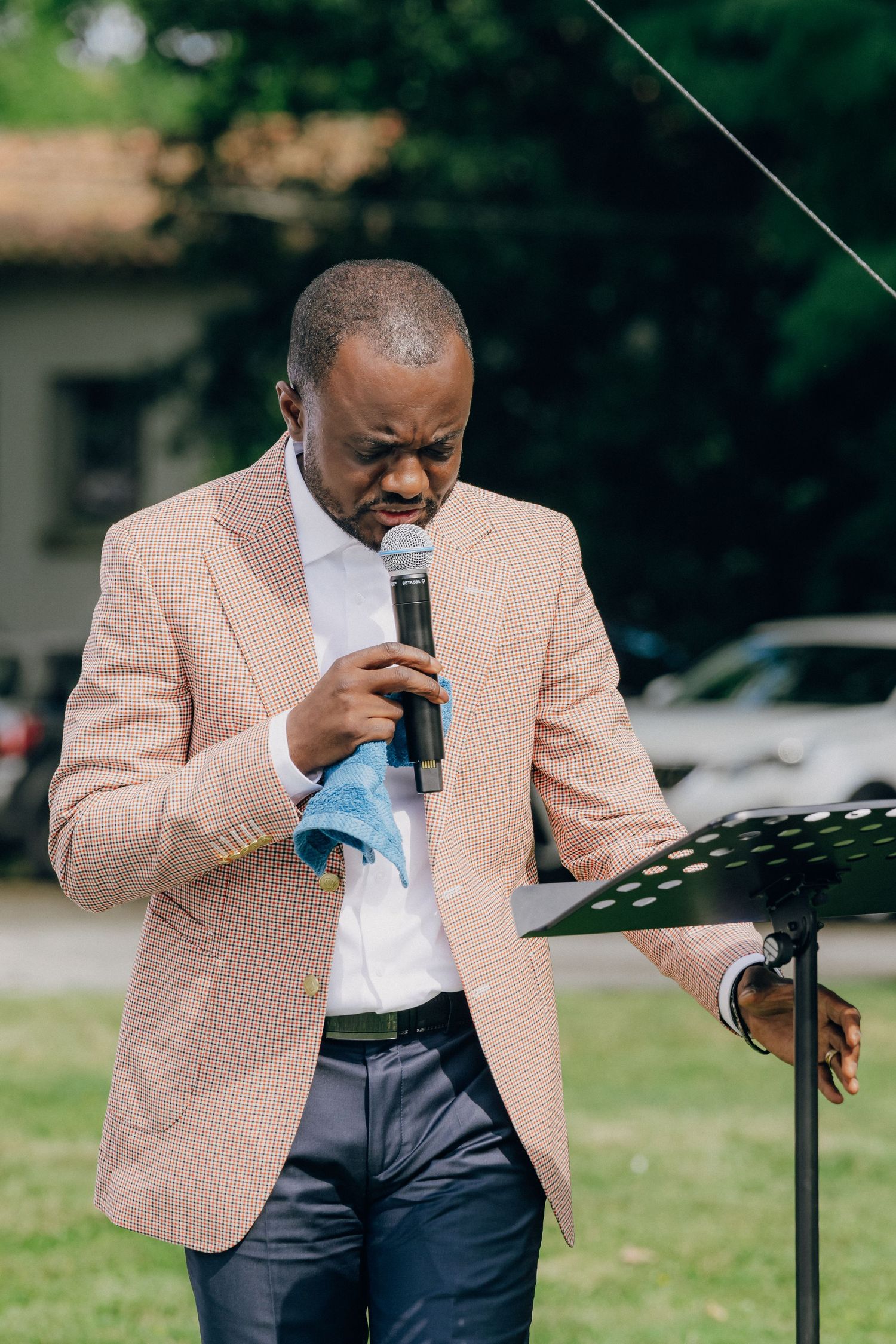 A person in a pink checkered blazer and white shirt reads from a document at a podium outdoors.