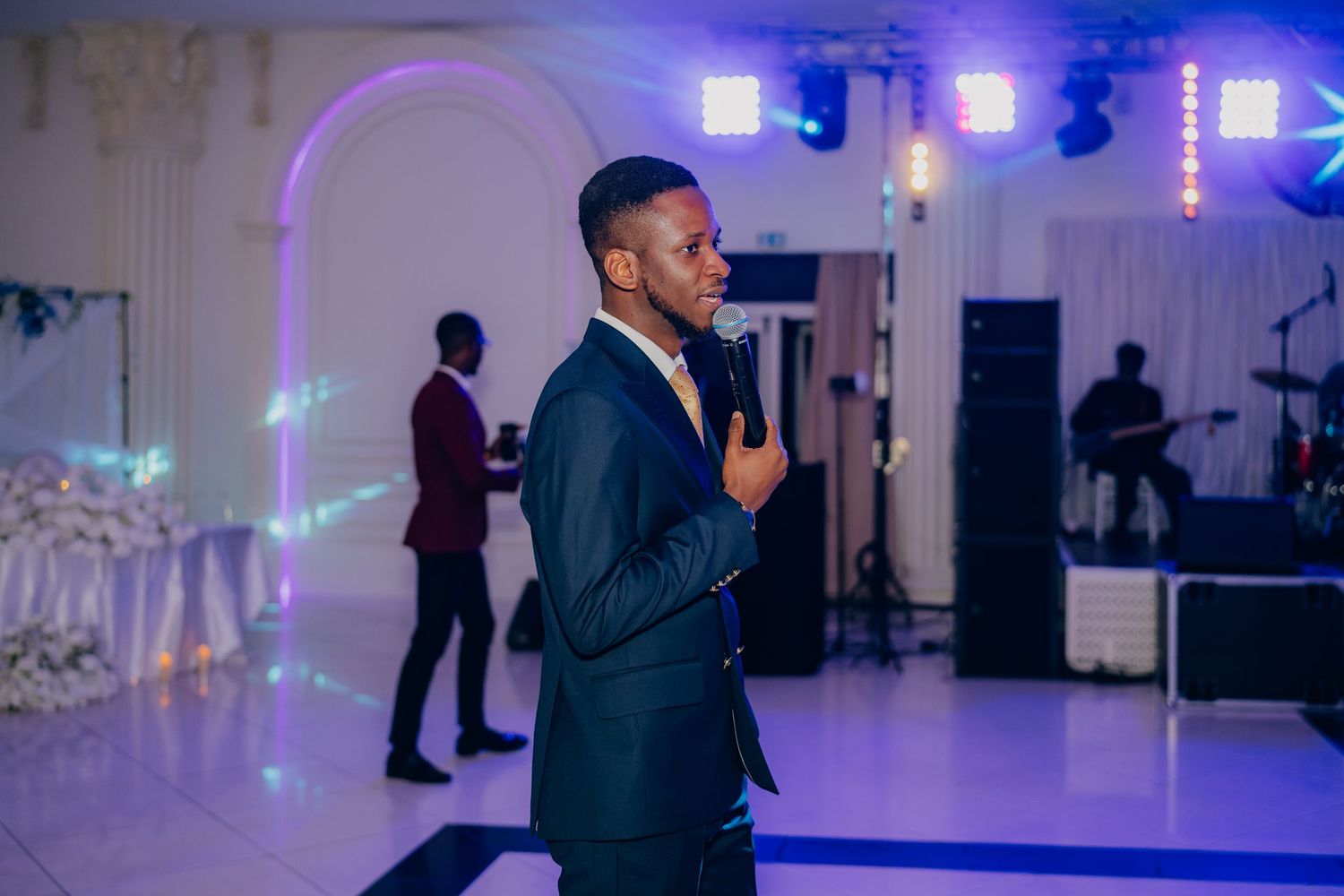 A performer in a navy suit speaks into a microphone at an elegant reception with purple lighting and arched decor.