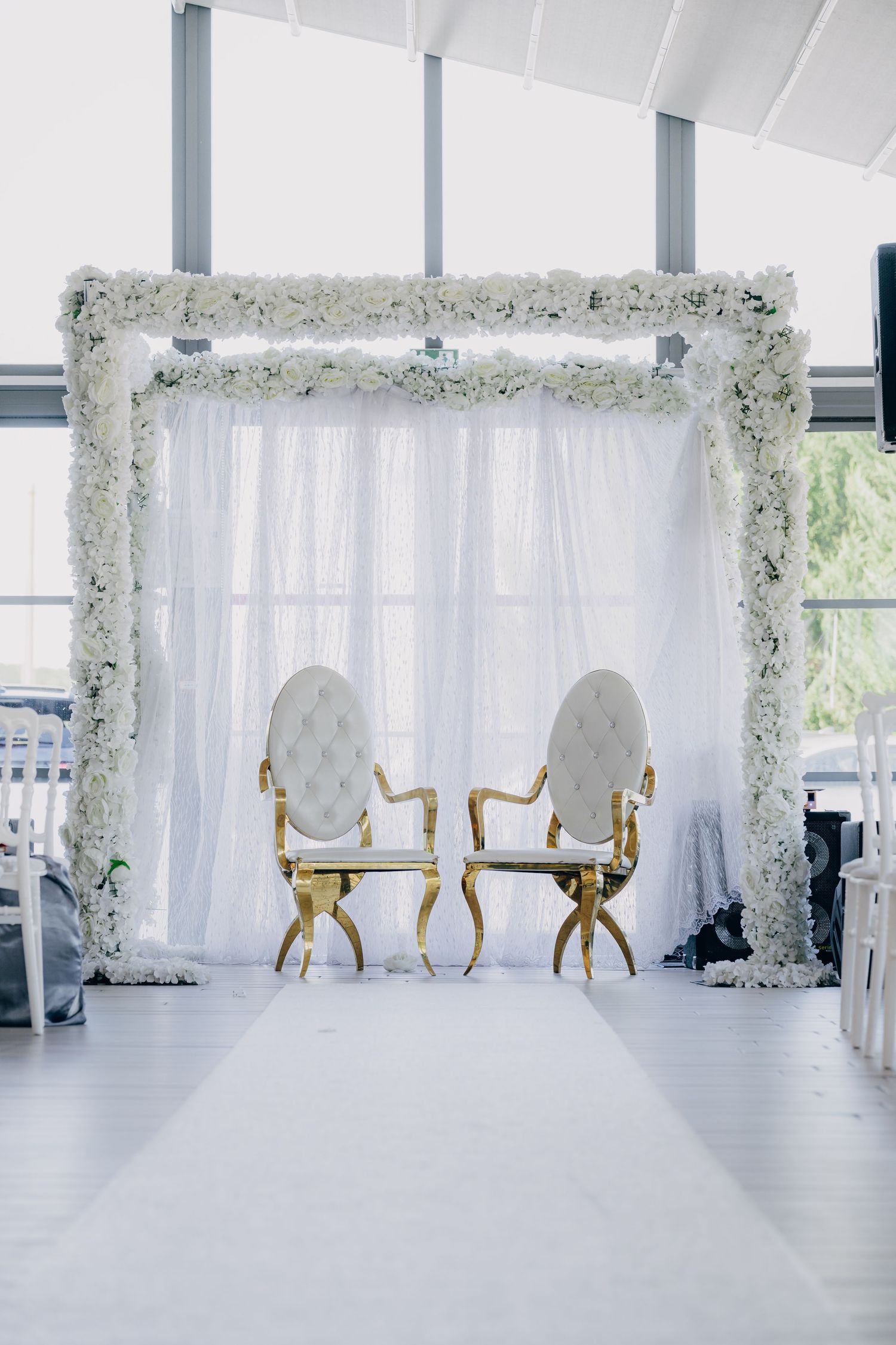 Elegant white wedding ceremony setup with floral arch and chairs in a bright modern venue with large windows.