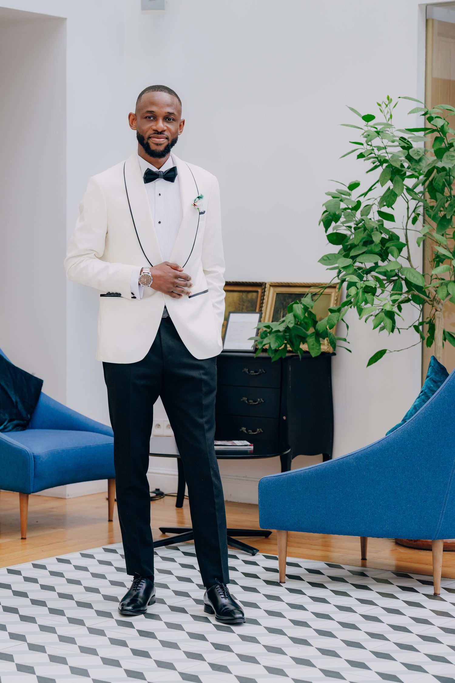 A person wearing a white tuxedo jacket and black bow tie stands on a patterned floor near blue chairs.
