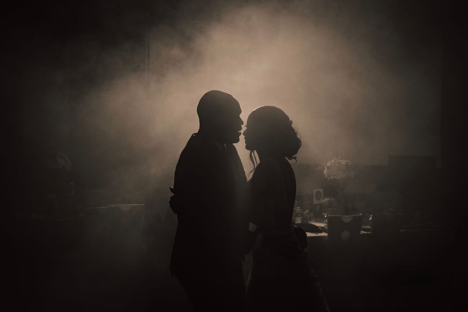 Two silhouettes stand close together in a moody, atmospheric scene with misty lighting and dark shadows.