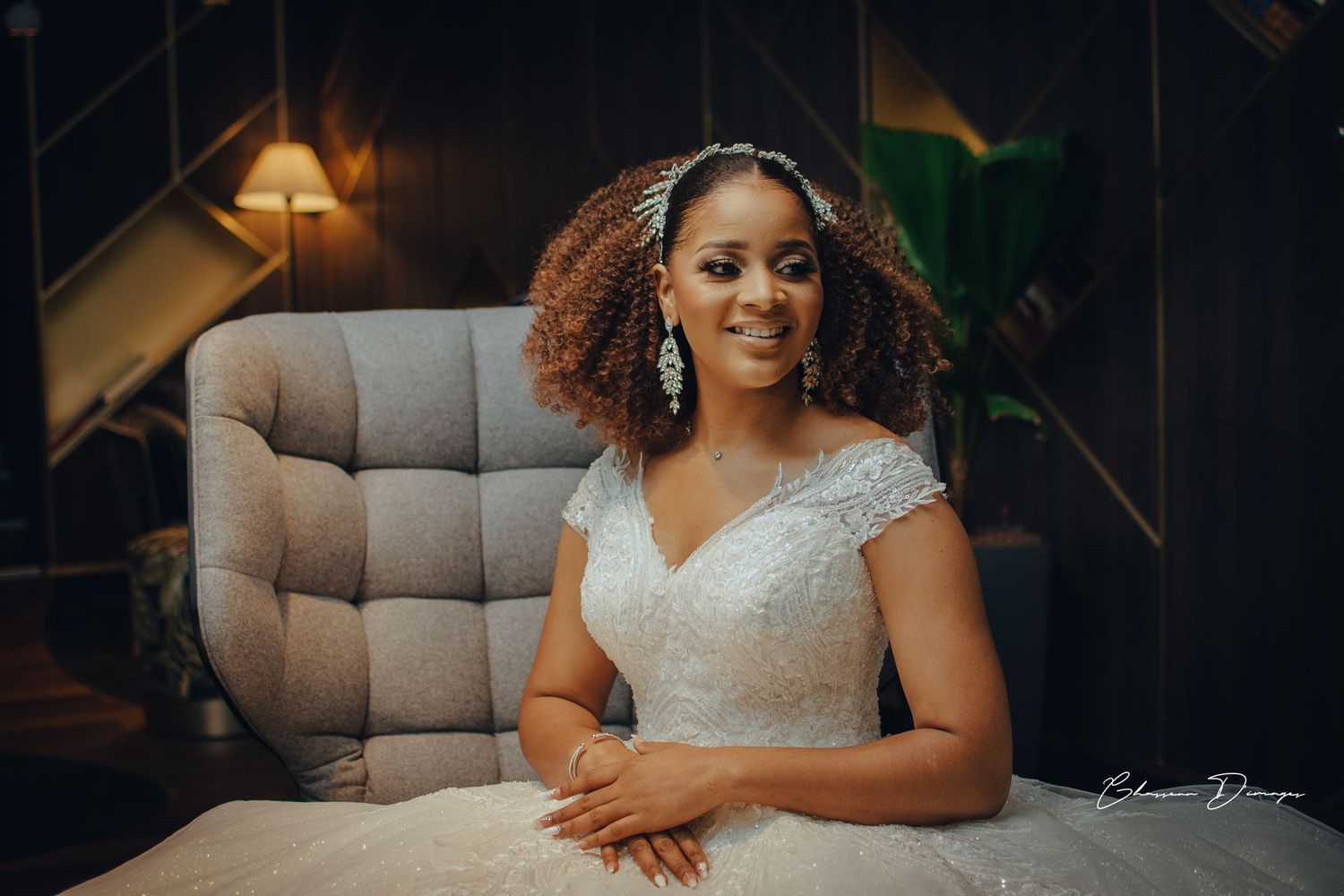 A bride in lace cap sleeve dress sits elegantly on beige chair.
