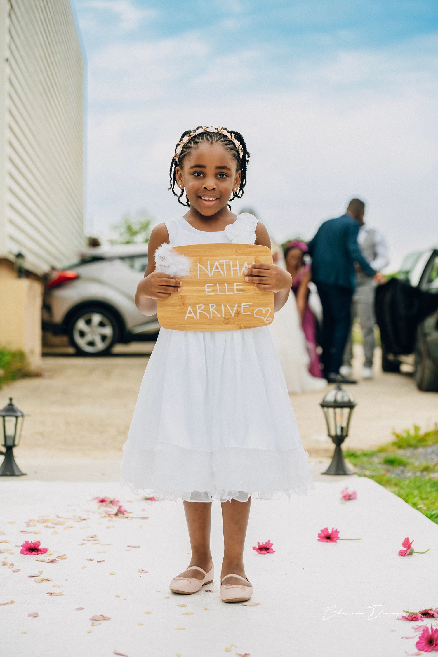 Young attendant in white dress holds wooden sign while standing on petal-strewn pathway.