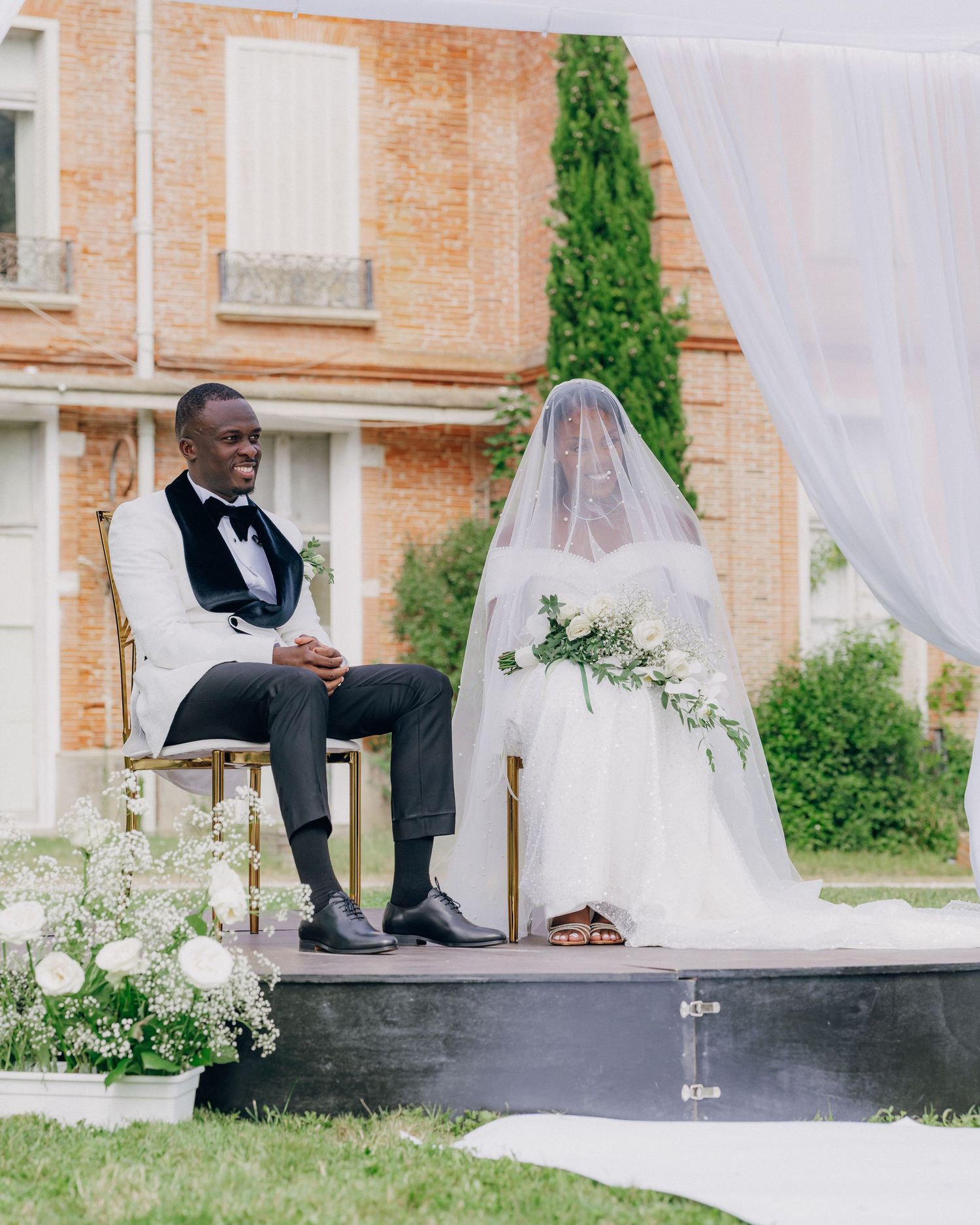 Elegant outdoor wedding ceremony setup with white draping and brick building backdrop.